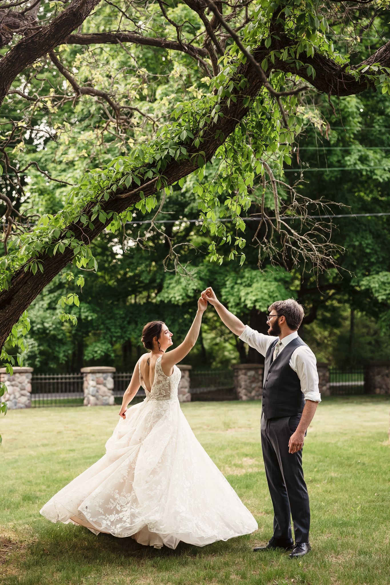 Couple dancing and twirling in elegant wedding attire under green tree canopy.
