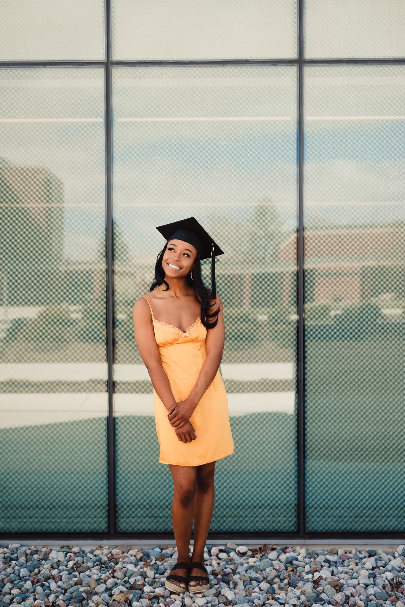 Graduate in orange dress poses on rooftop with modern glass building backdrop for graduation photos.