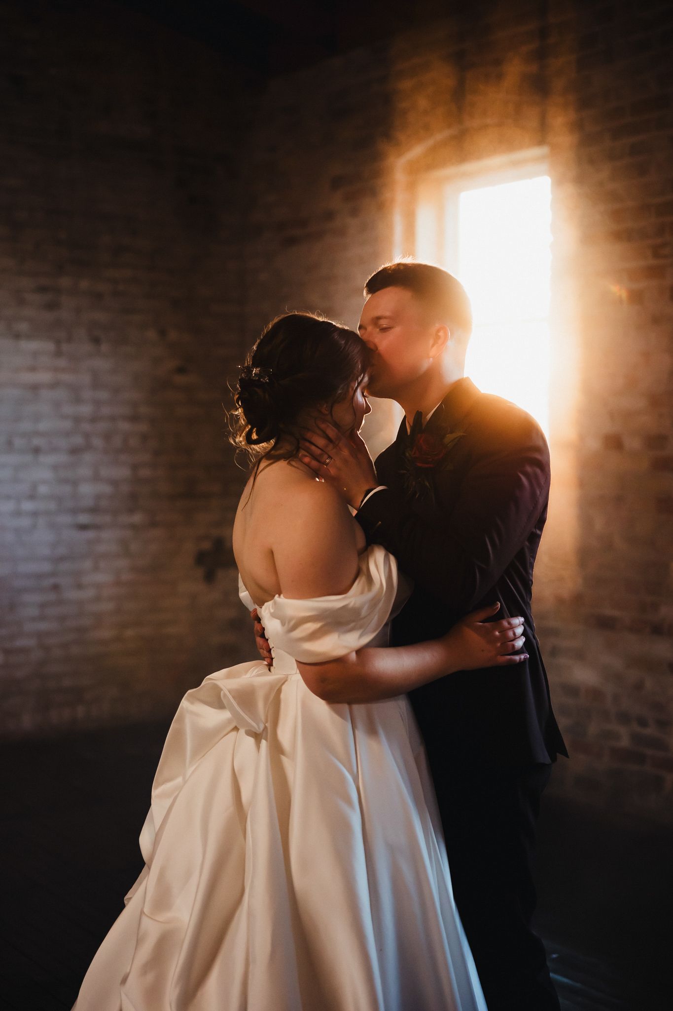 A backlit romantic moment in a rustic brick room with dramatic window lighting.