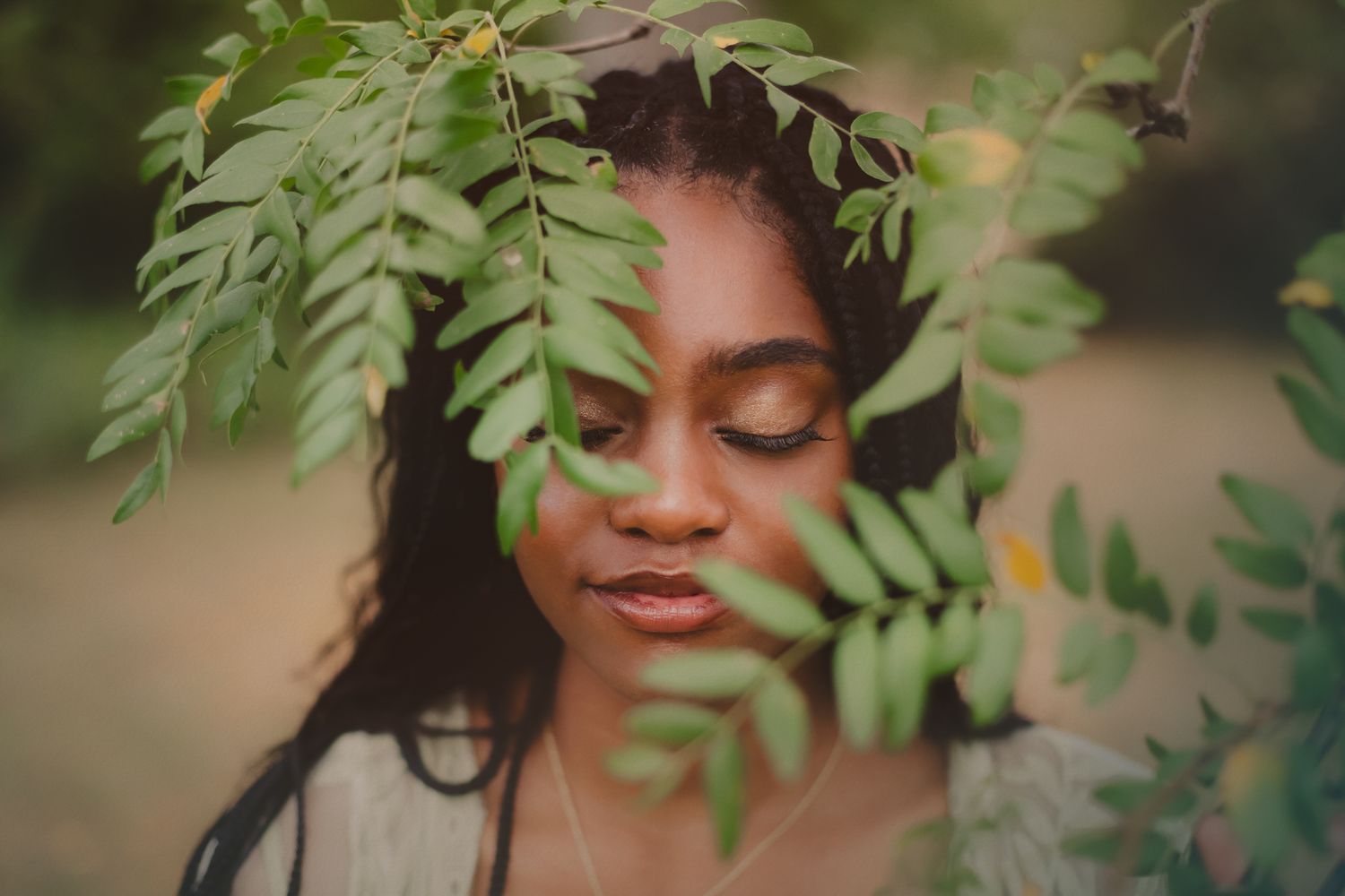 A person poses in nature surrounded by lush green leaves and foliage in soft natural lighting.