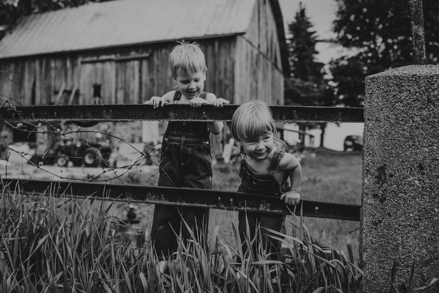 A black and white photograph shows two children leaning on a fence post near a rustic barn in a rural setting.