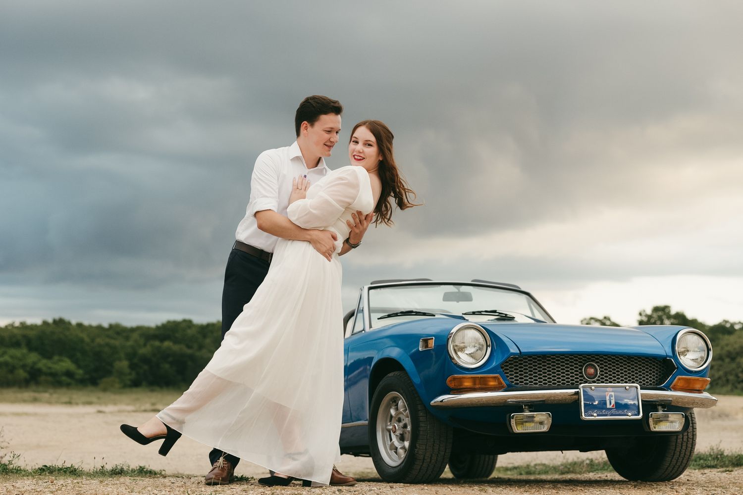 Romantic portraits with a classic blue vintage car under stormy skies.