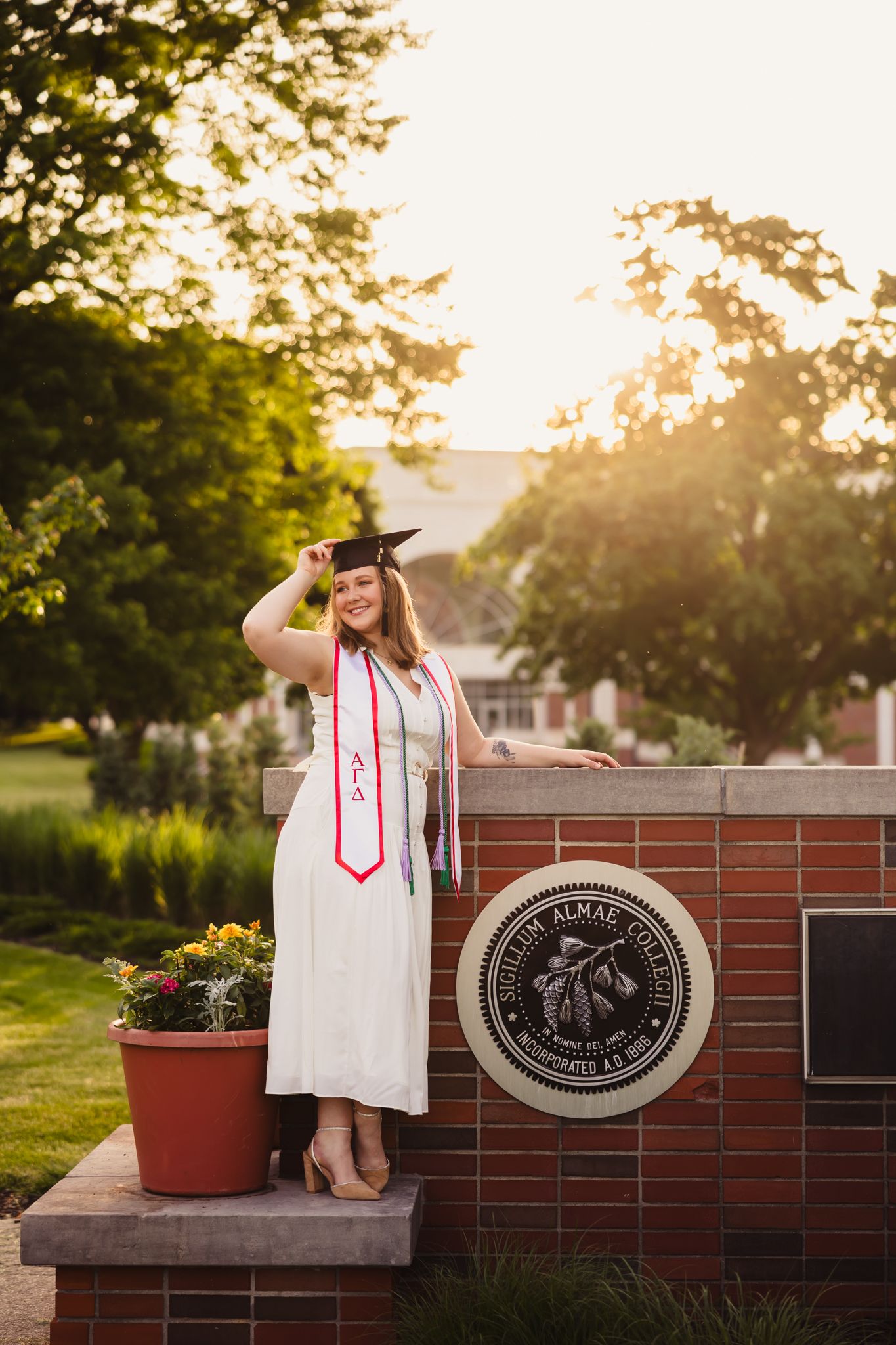 Graduate in white cap and gown sits on brick wall with university seal at golden hour.