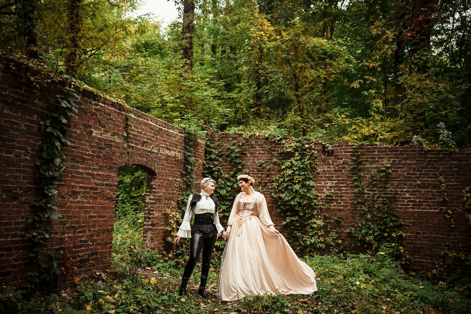 A couple walks along a garden path bordered by brick walls and lush greenery during a wedding photoshoot.