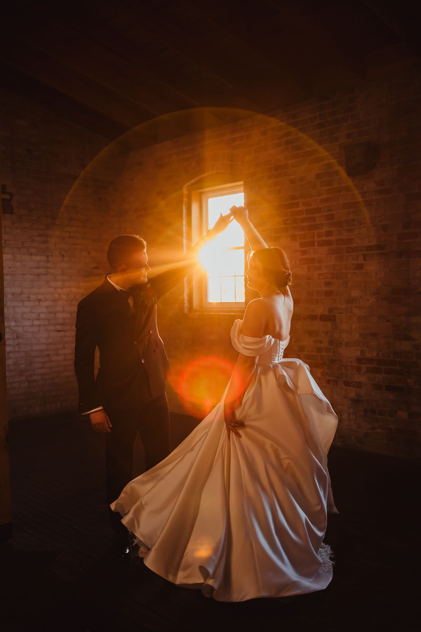 A romantic wedding couple silhouetted against sunlight streaming through a brick window, with the bride in a flowing white gown.