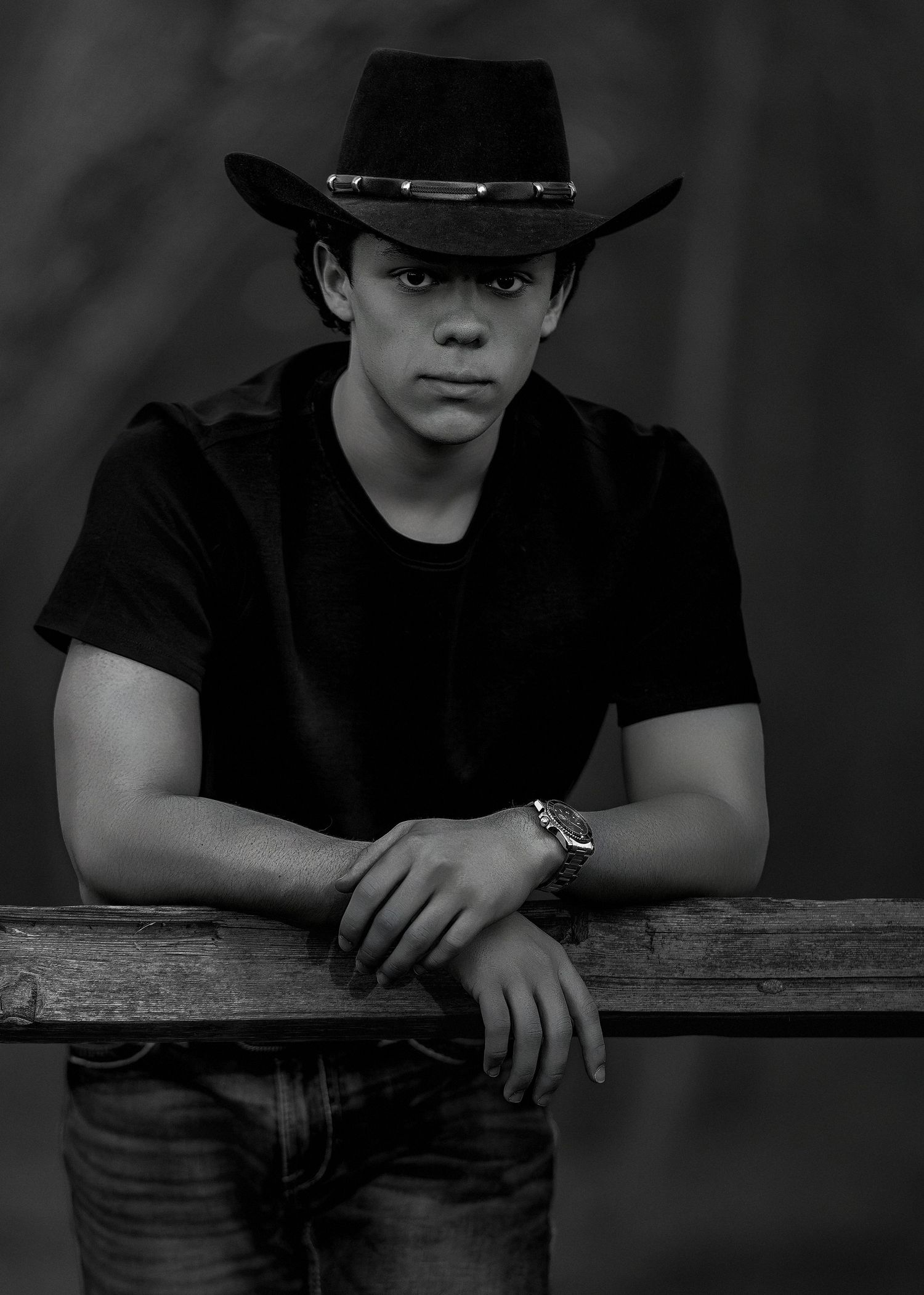 Black and white portrait of someone wearing a black cowboy hat and t-shirt leaning against a wooden railing.