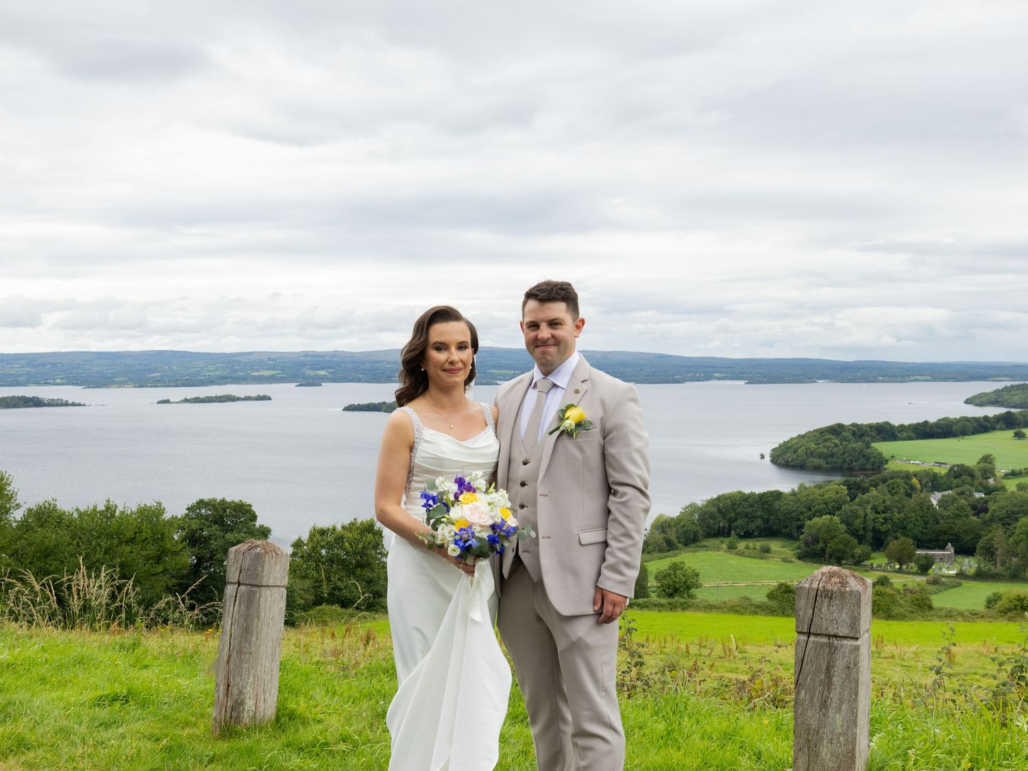 A couple poses on a hilltop overlooking a scenic lake and green landscape on their wedding day.