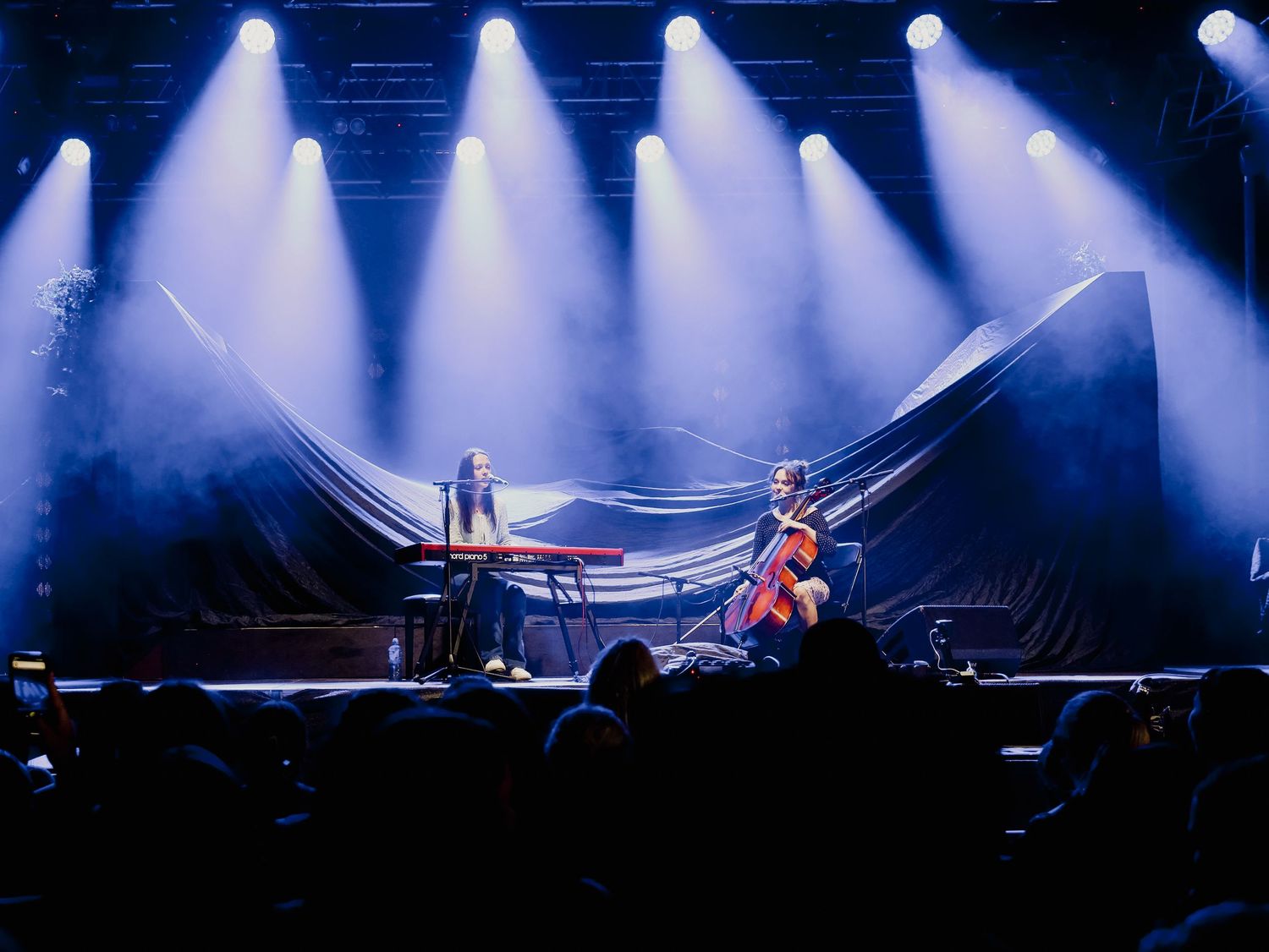 A dramatic concert stage illuminated by bright blue spotlights with silhouetted performers and equipment visible.
