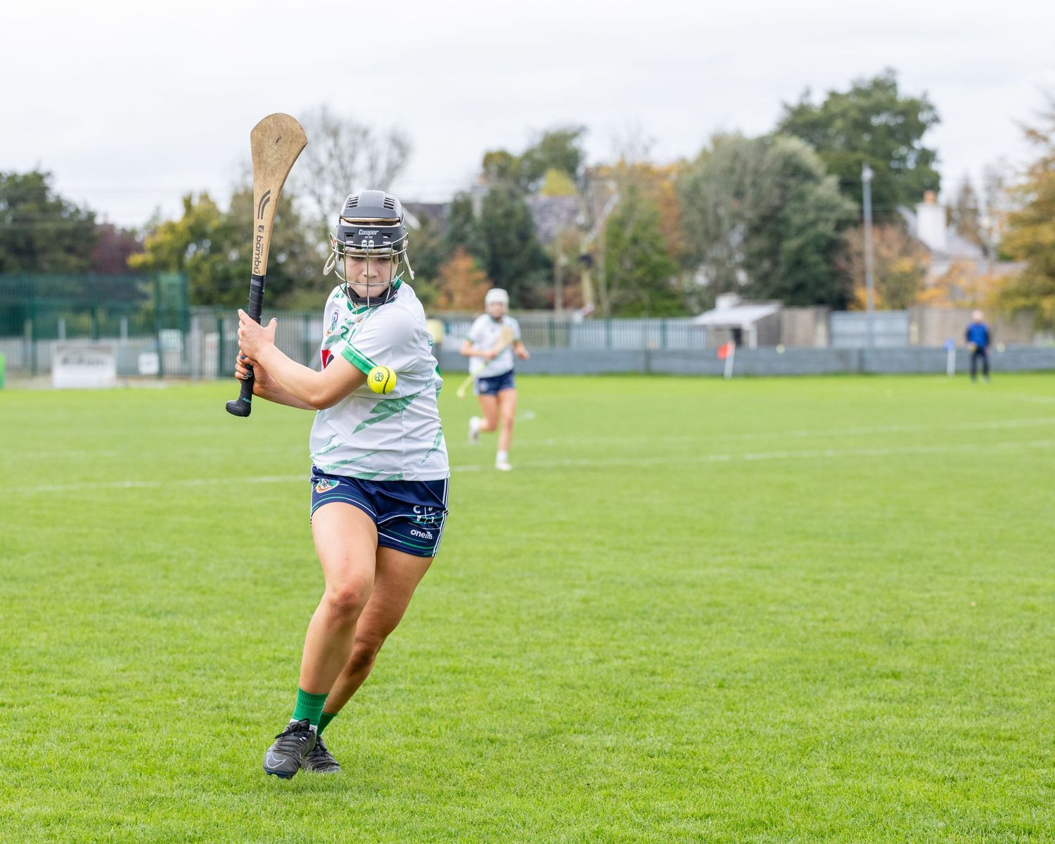 Camogie on a green grass field during a match with one player preparing to strike the ball.