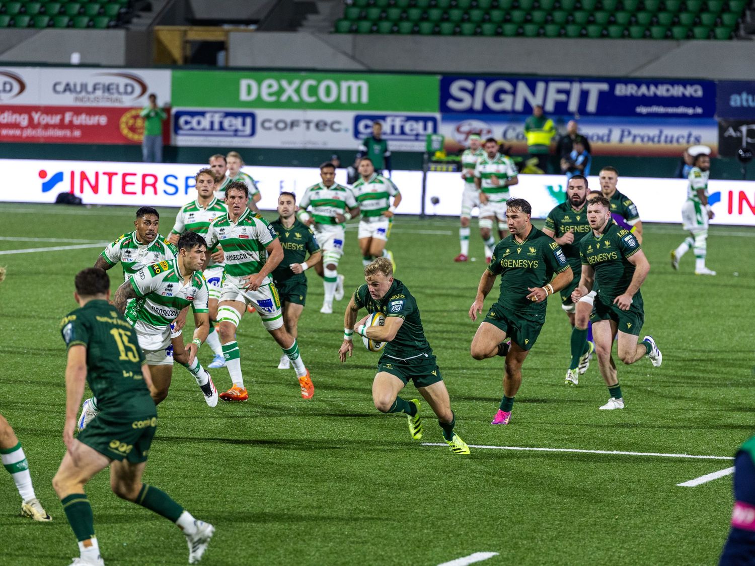 Rugby players compete for possession during an intense match on a green field with stadium advertising boards visible.