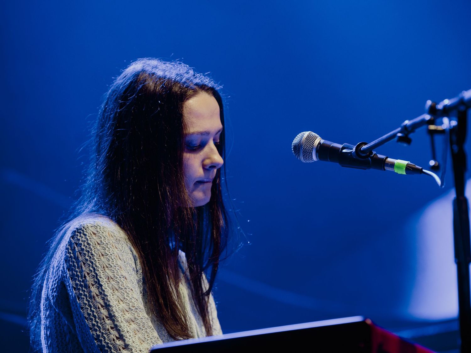 Singer performs on stage with piano in dark blue lighting during live concert performance.
