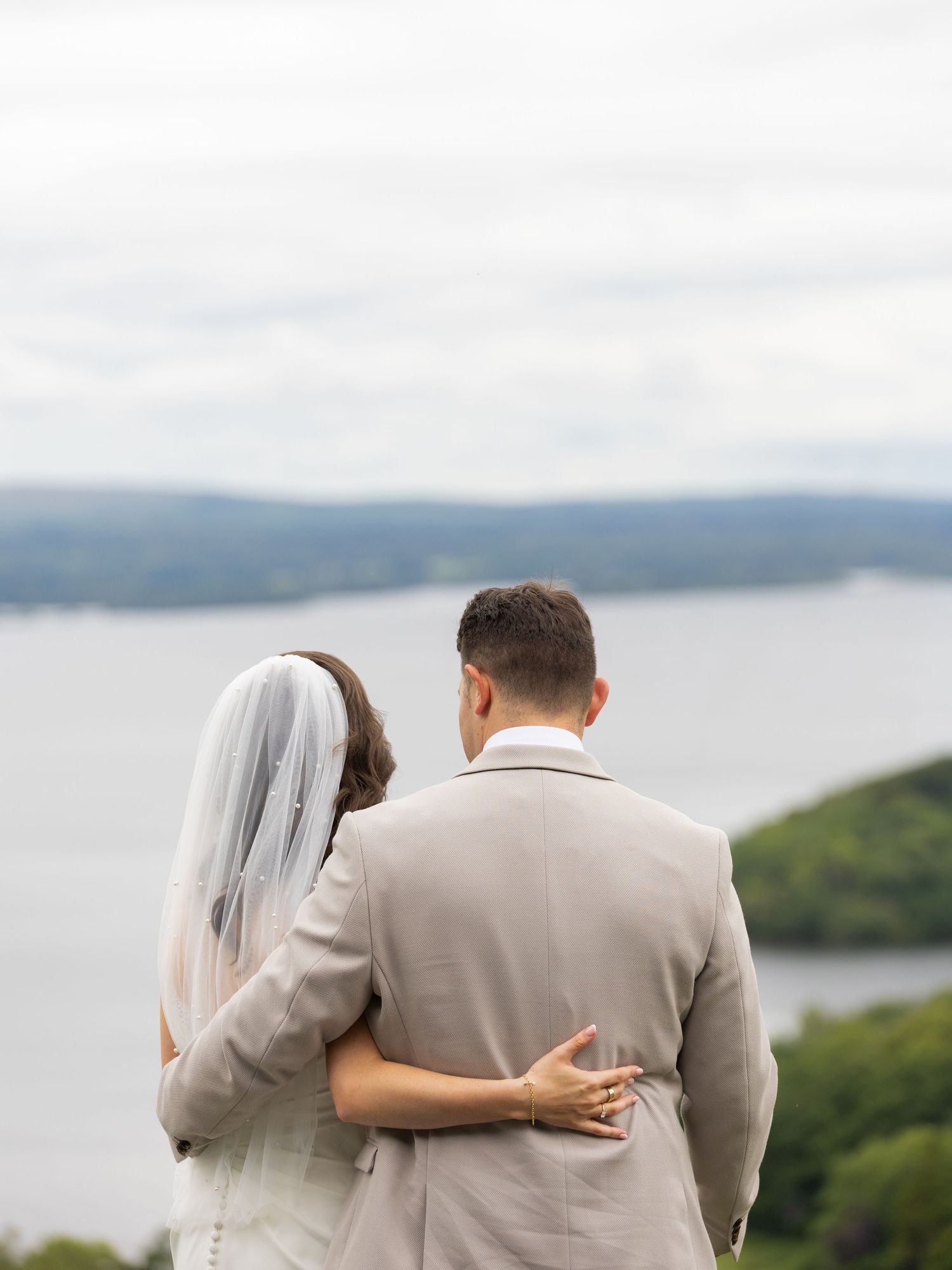 Newlyweds embrace while admiring a breathtaking lake view from a hilltop vantage point.