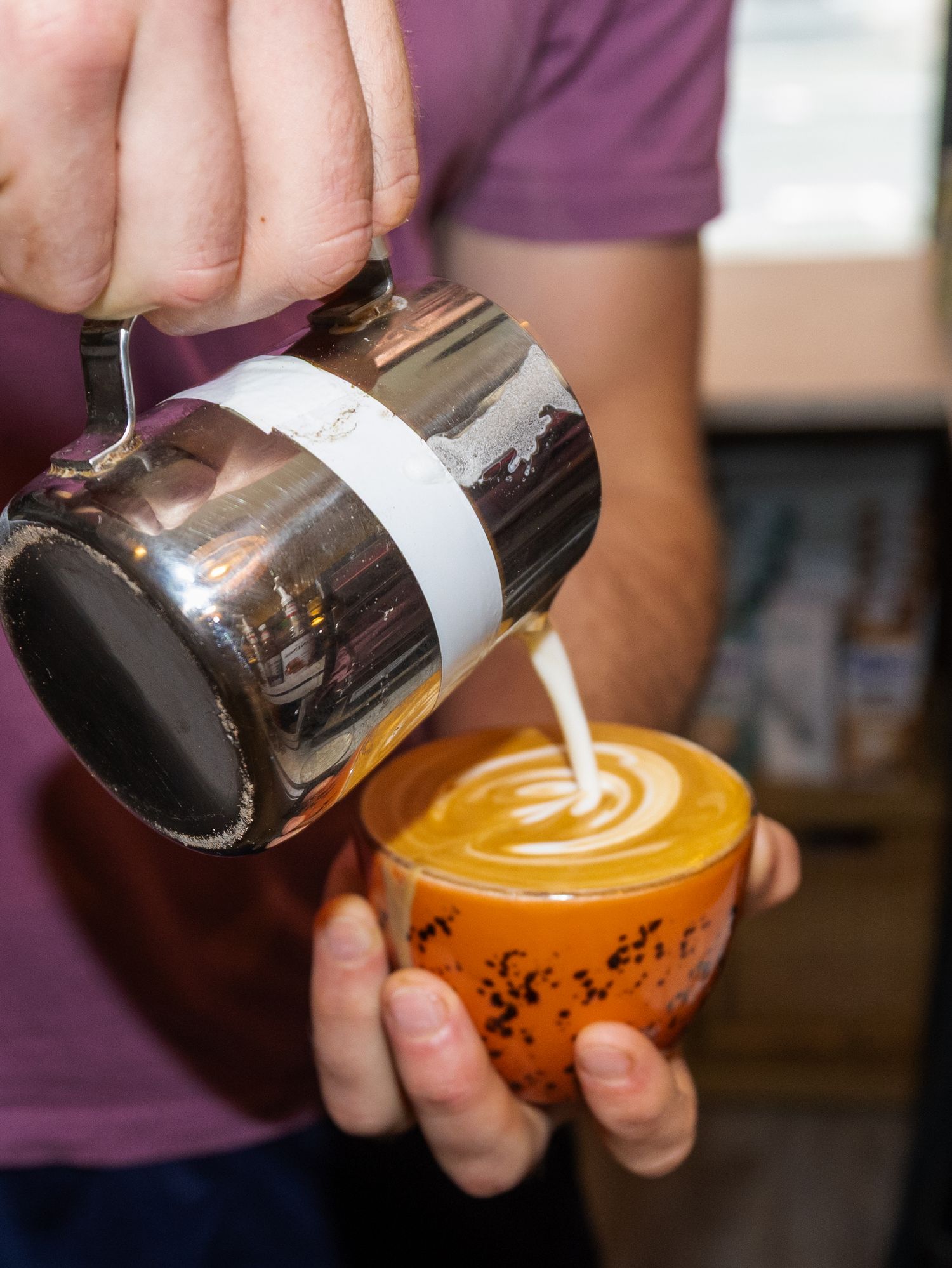 Barista pouring steamed milk into an orange coffee cup creating latte art.
