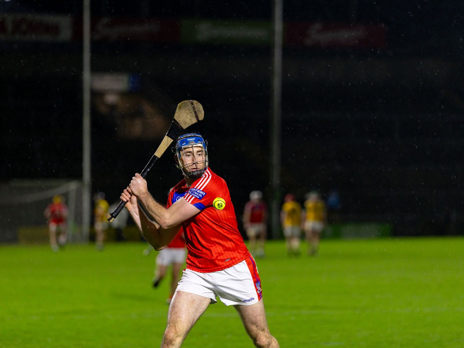Hurling player in red and white jersey makes a striking motion with their hurley stick during a nighttime match.