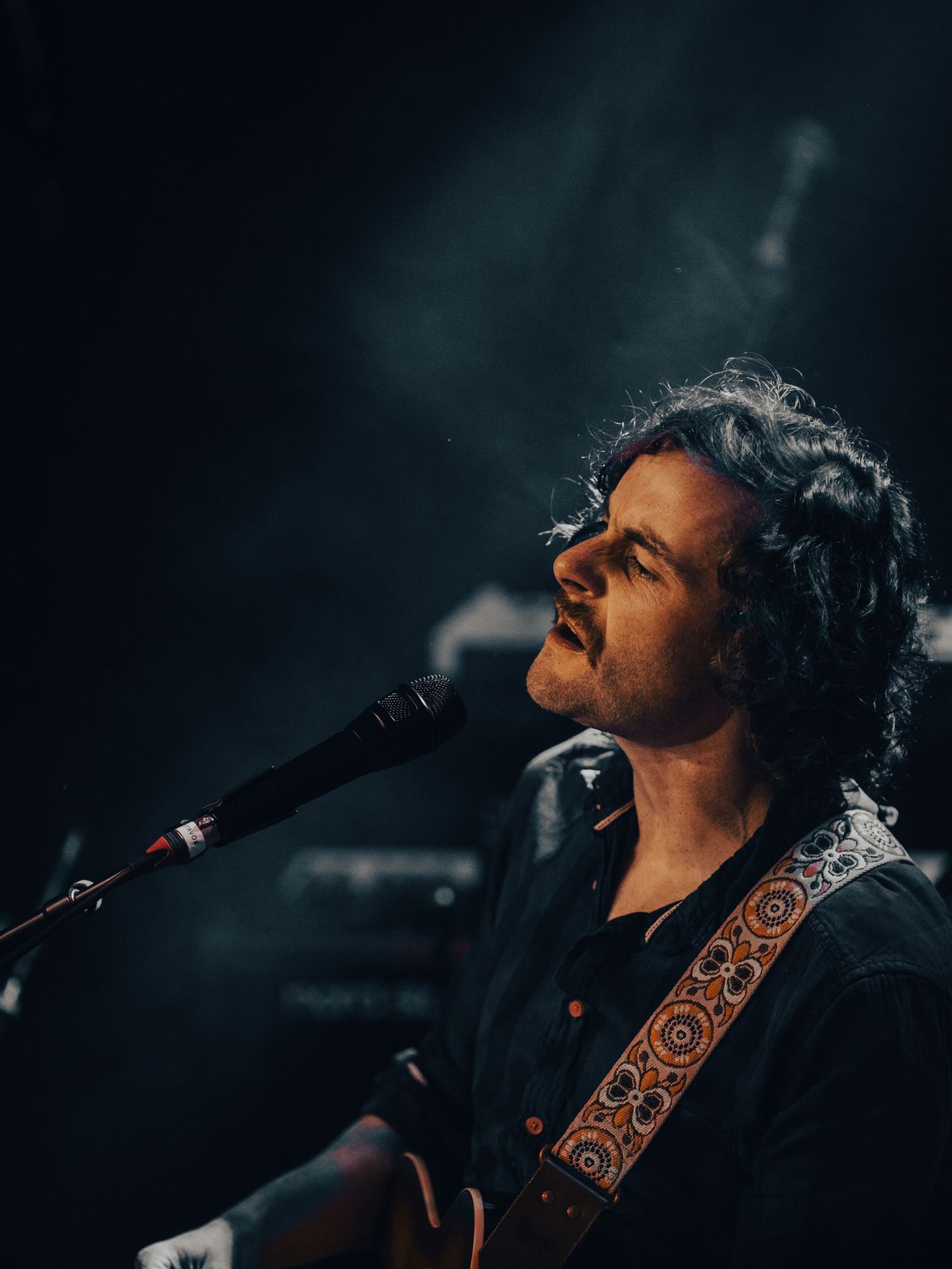 Singer performing with acoustic guitar against dark moody stage lighting.