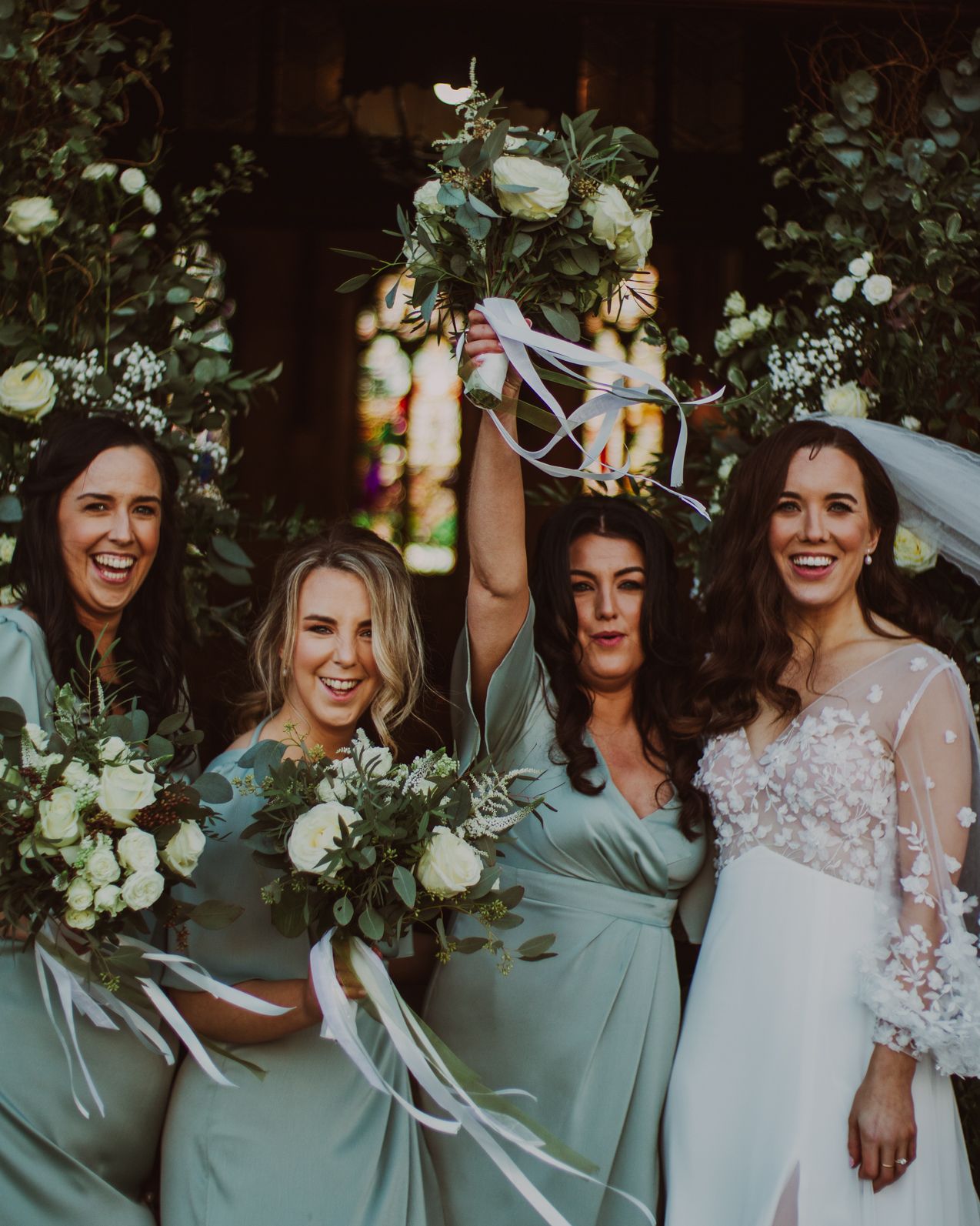 A group of bridesmaids in sage green dresses pose together with the bride in a lace gown during a wedding celebration.