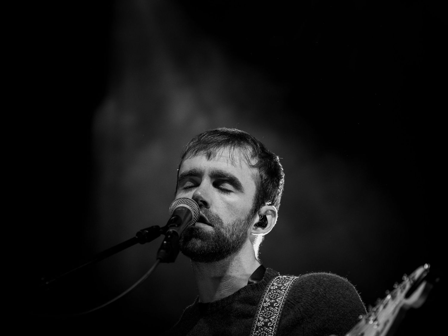 A dramatic black and white performance photo of a musician singing into a microphone on stage.