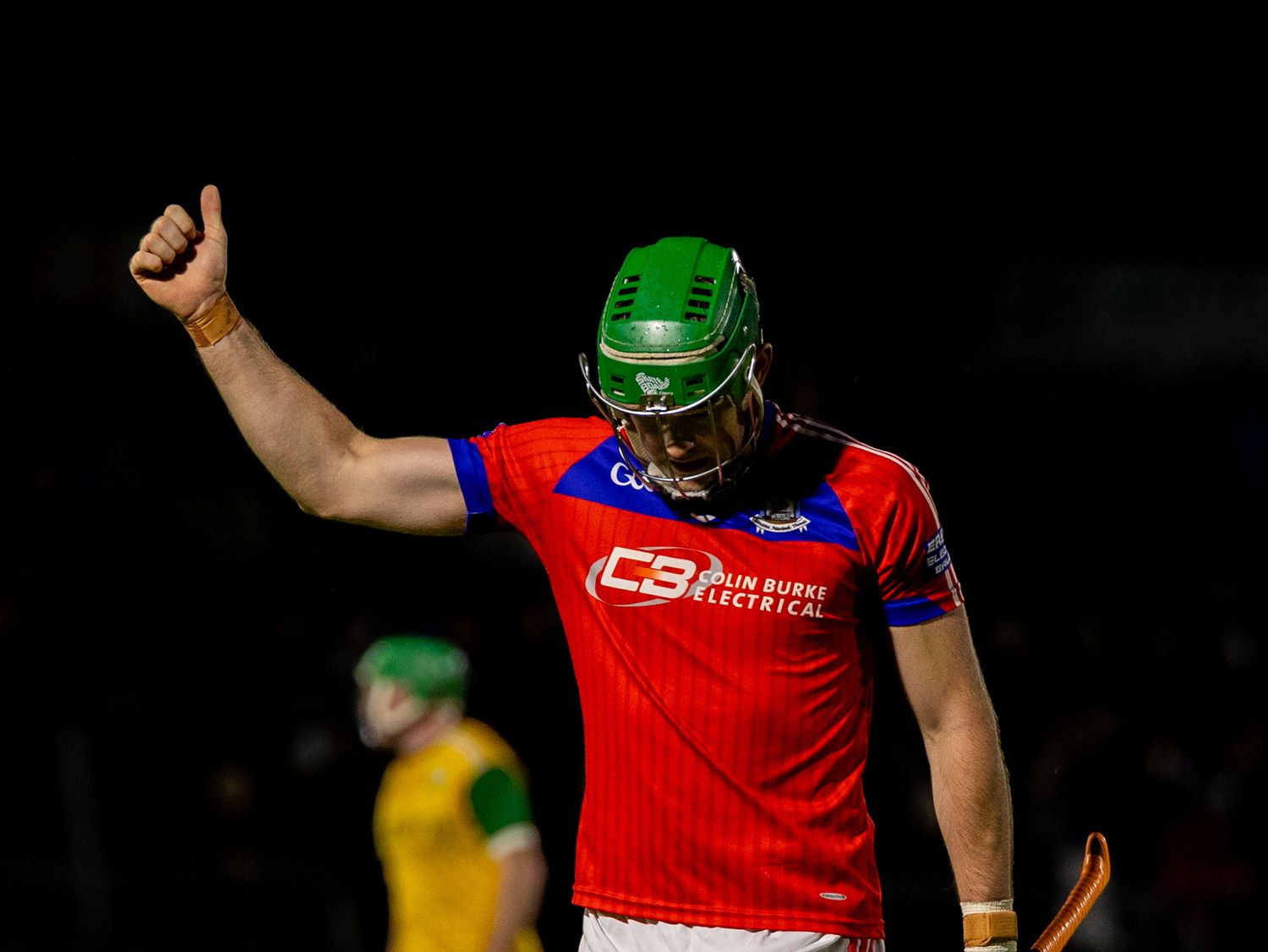 Cricket batsman in red jersey and green helmet plays a shot during a match under stadium lights.