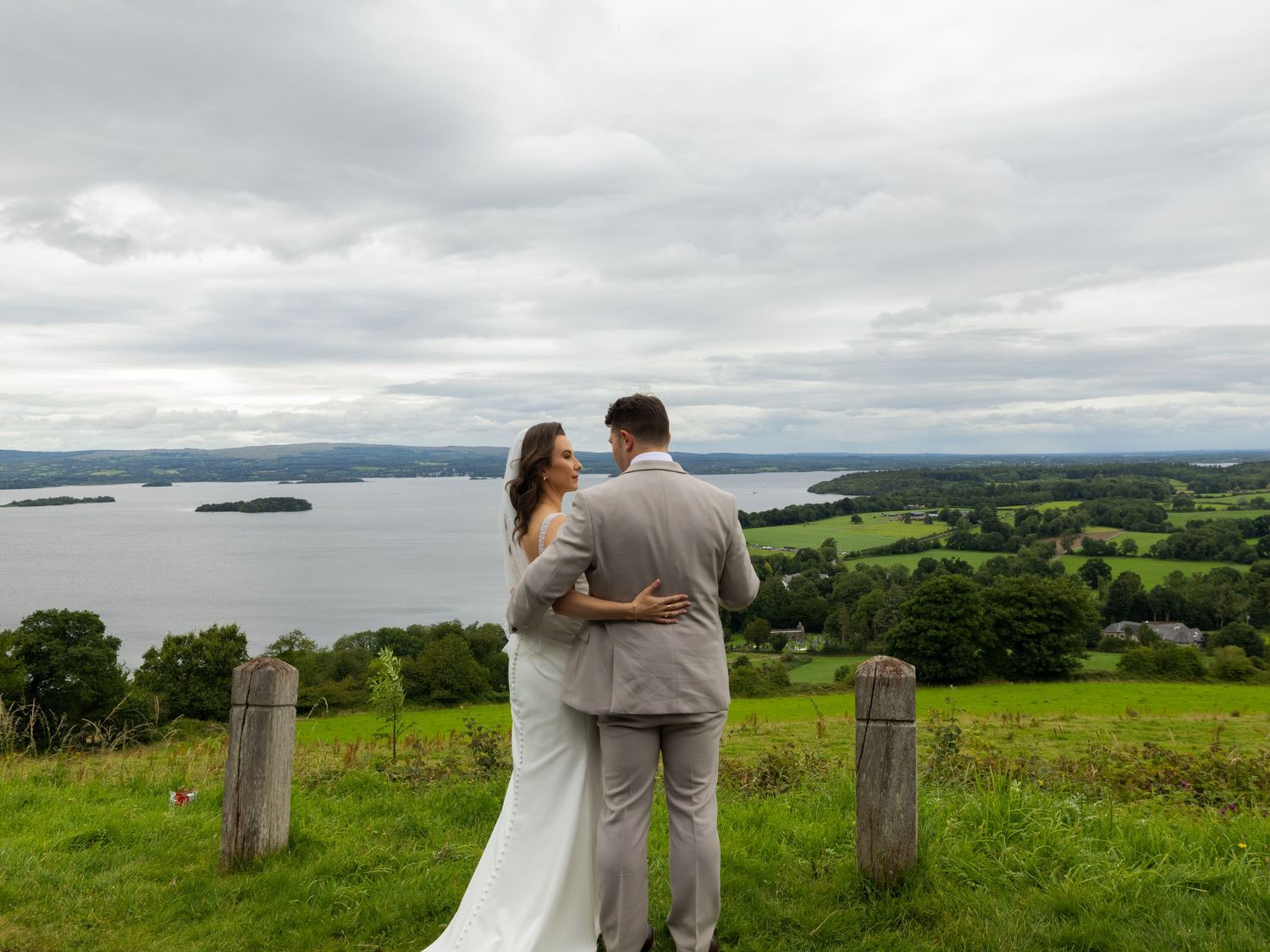 A couple stands together overlooking a scenic coastal landscape with rolling green hills and distant islands.