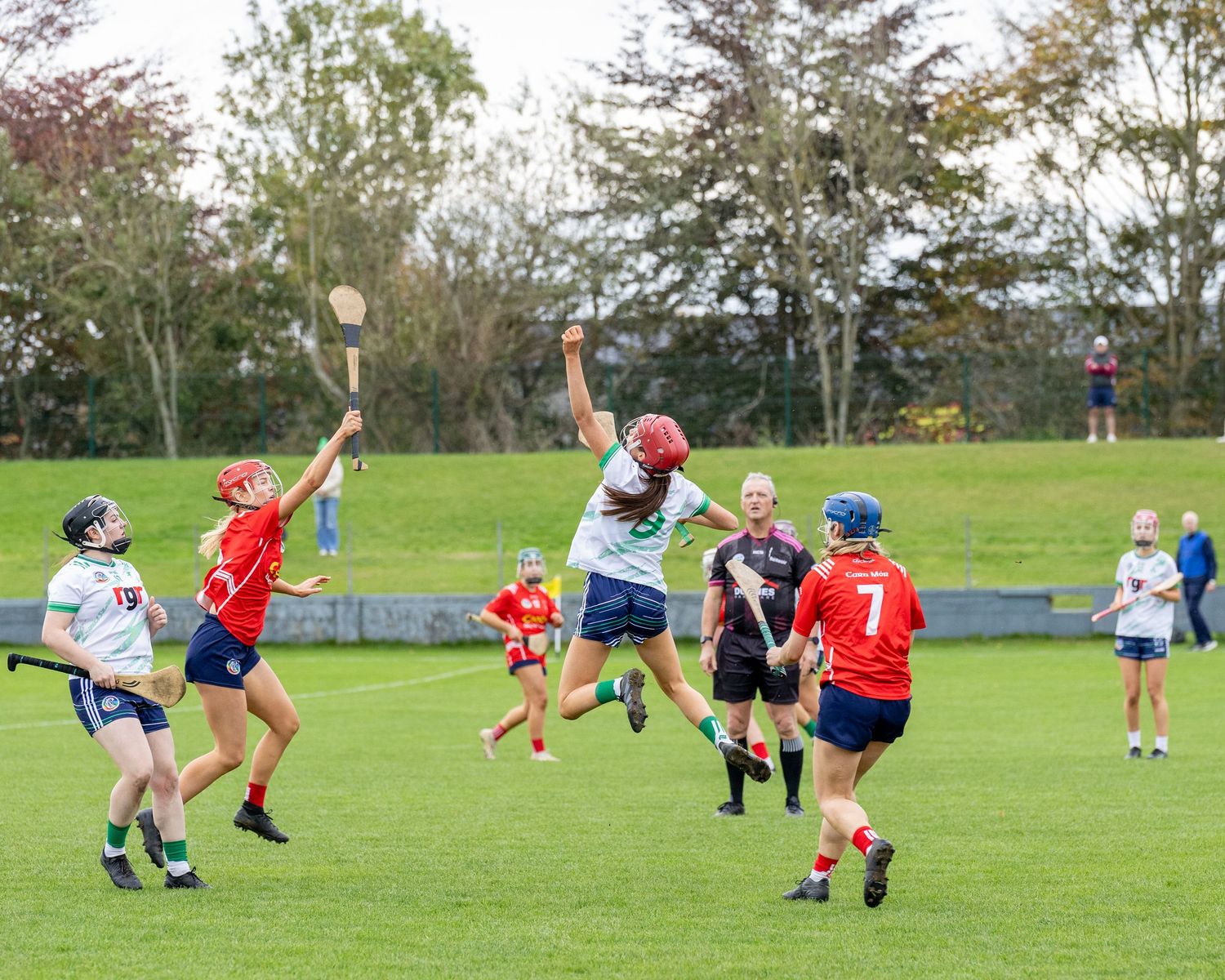 Players jump for the ball during an outdoor hurling match on a grass field surrounded by trees.