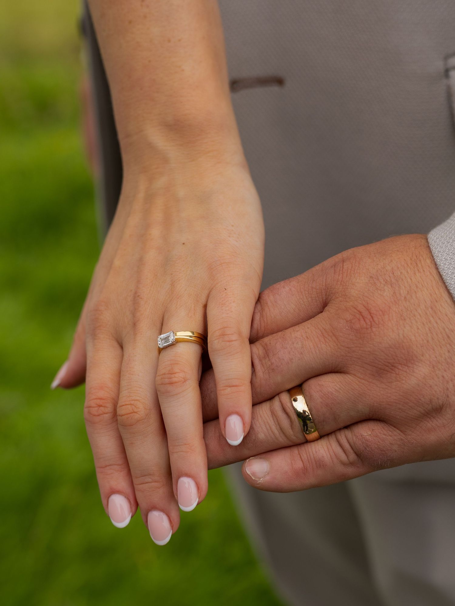 Close-up view of hands wearing wedding rings holding each other against a blurred green outdoor background.