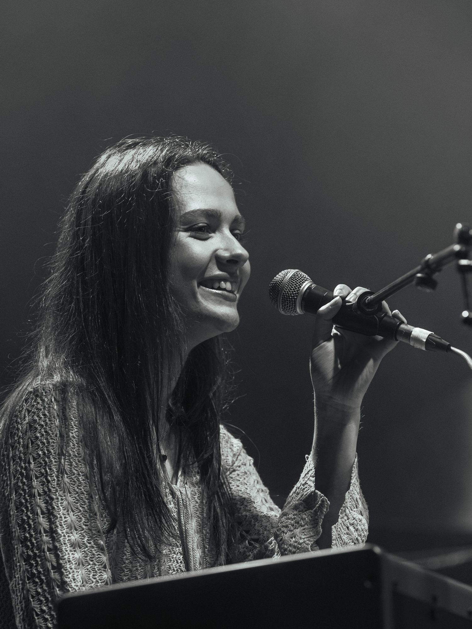 Singer with long dark hair performs passionately at a microphone in dramatic black and white lighting.