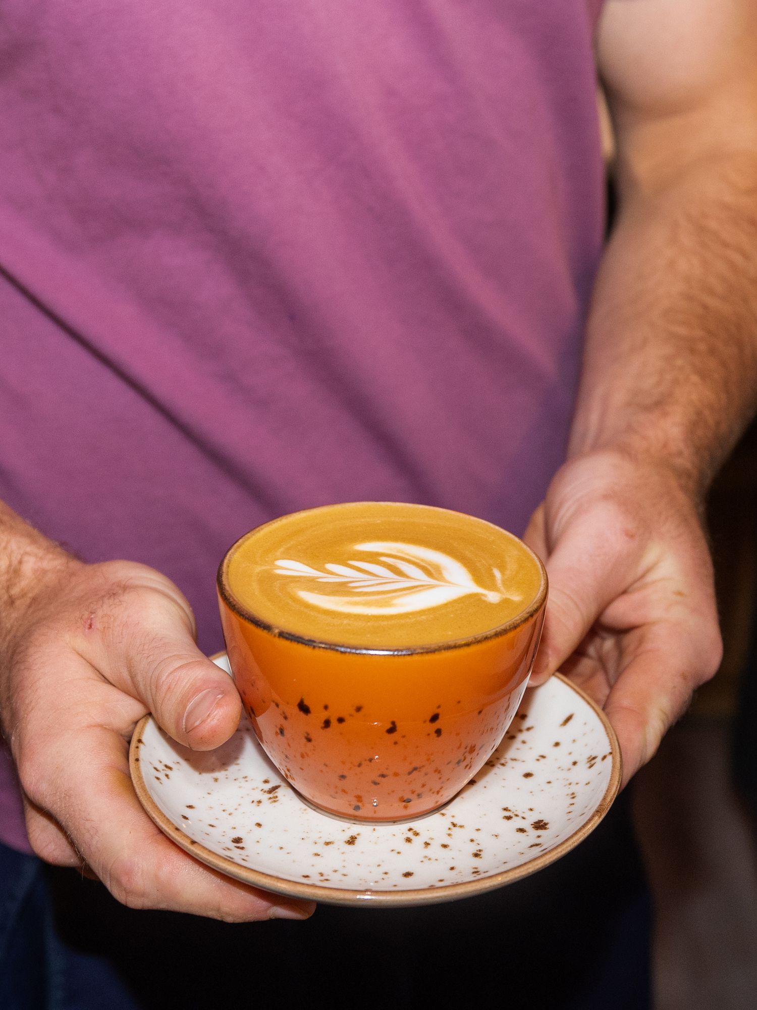 Hands holding orange ceramic cups with latte art designs on white saucers against a purple background.