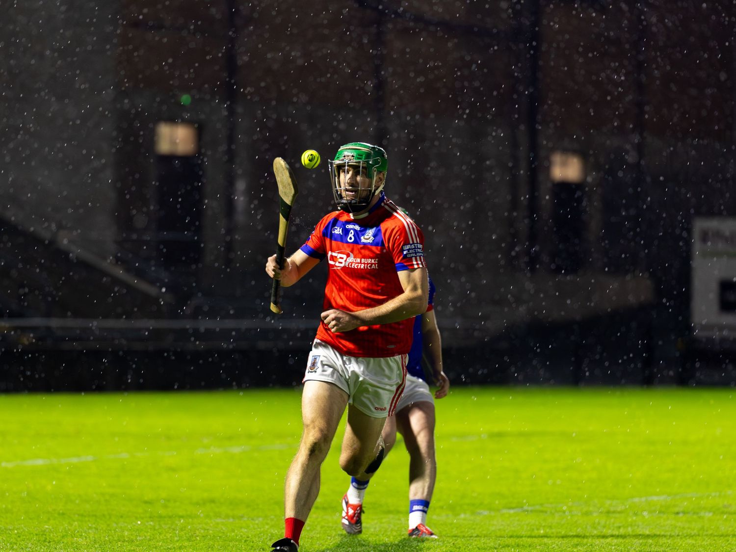A hurling player in red and white uniform swings their stick during a nighttime match under stadium lights.