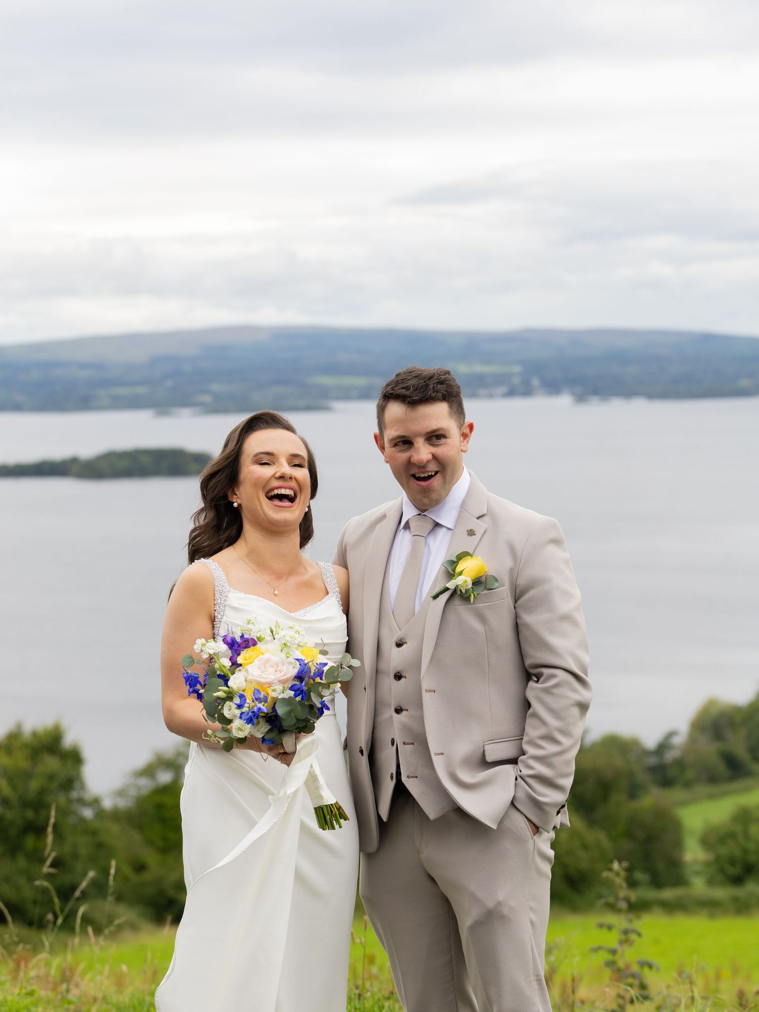Wedding couple laughs together holding colorful bouquet by lakeside overlook.