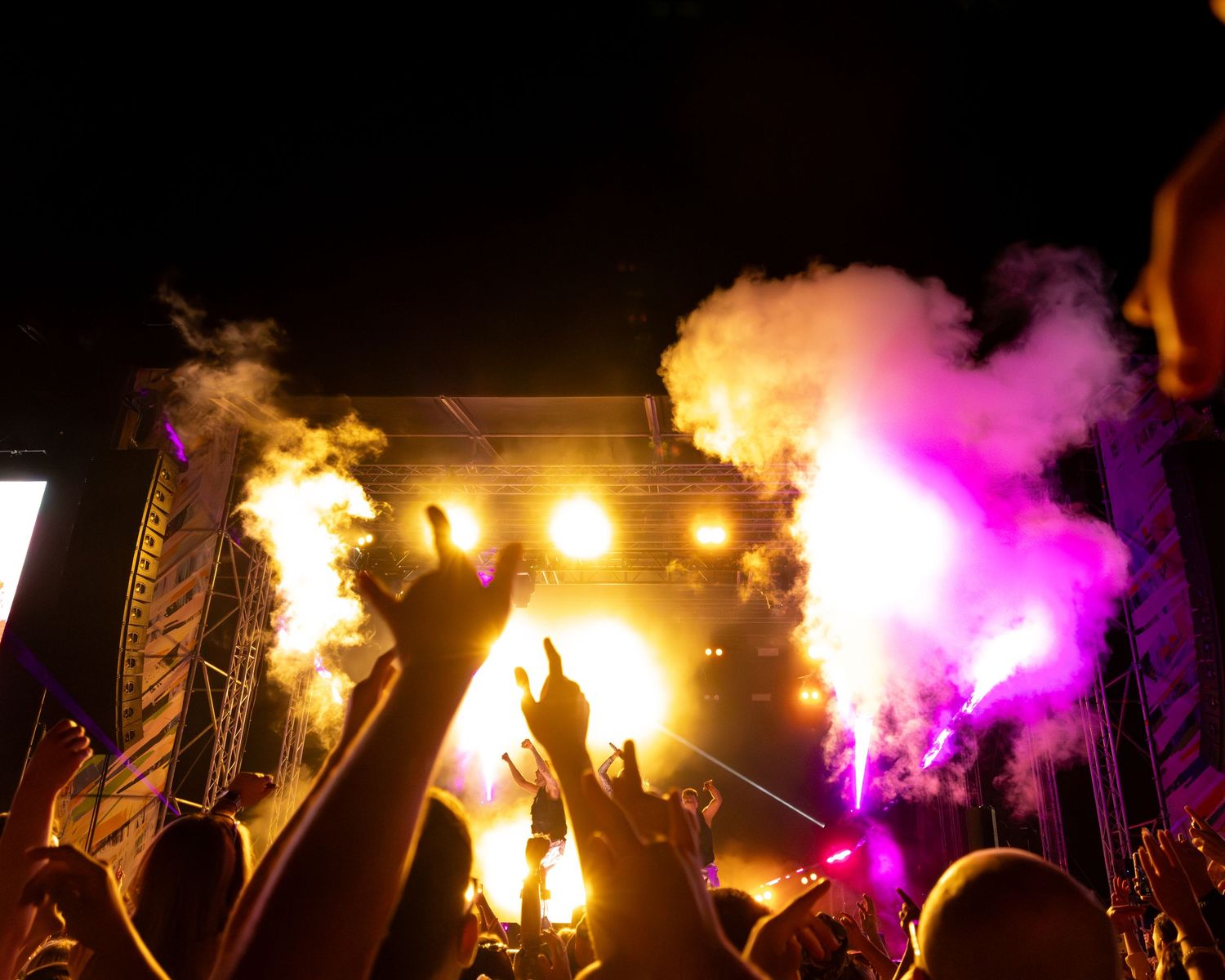 Dramatic concert stage with colorful pyrotechnics and smoke effects as crowd raises hands in silhouette.