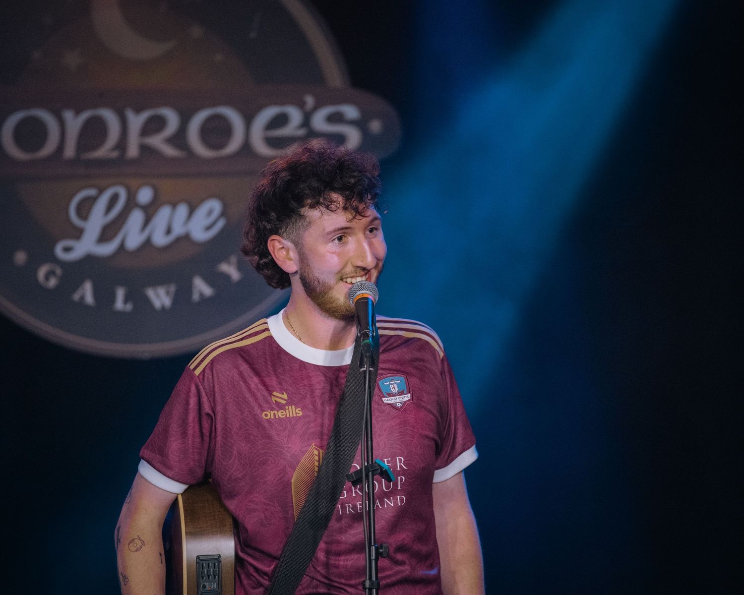 A performer in a maroon sports jersey stands at a microphone on stage with blue lighting at Monroe's venue.