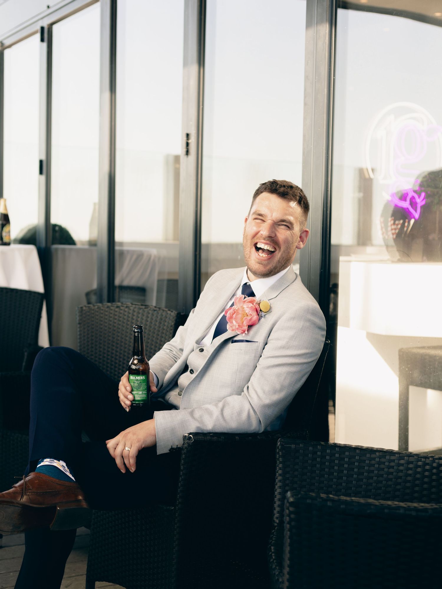 Wedding party member relaxes on outdoor patio furniture in white suit while holding a beverage.