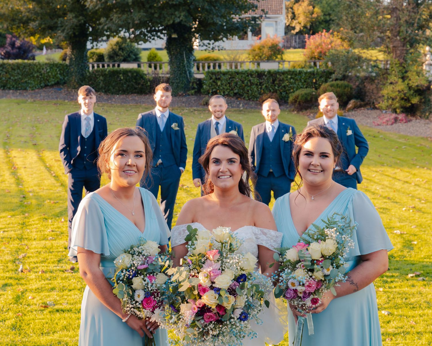 Bridal party in pale blue dresses poses with bouquets against golden sunset field.