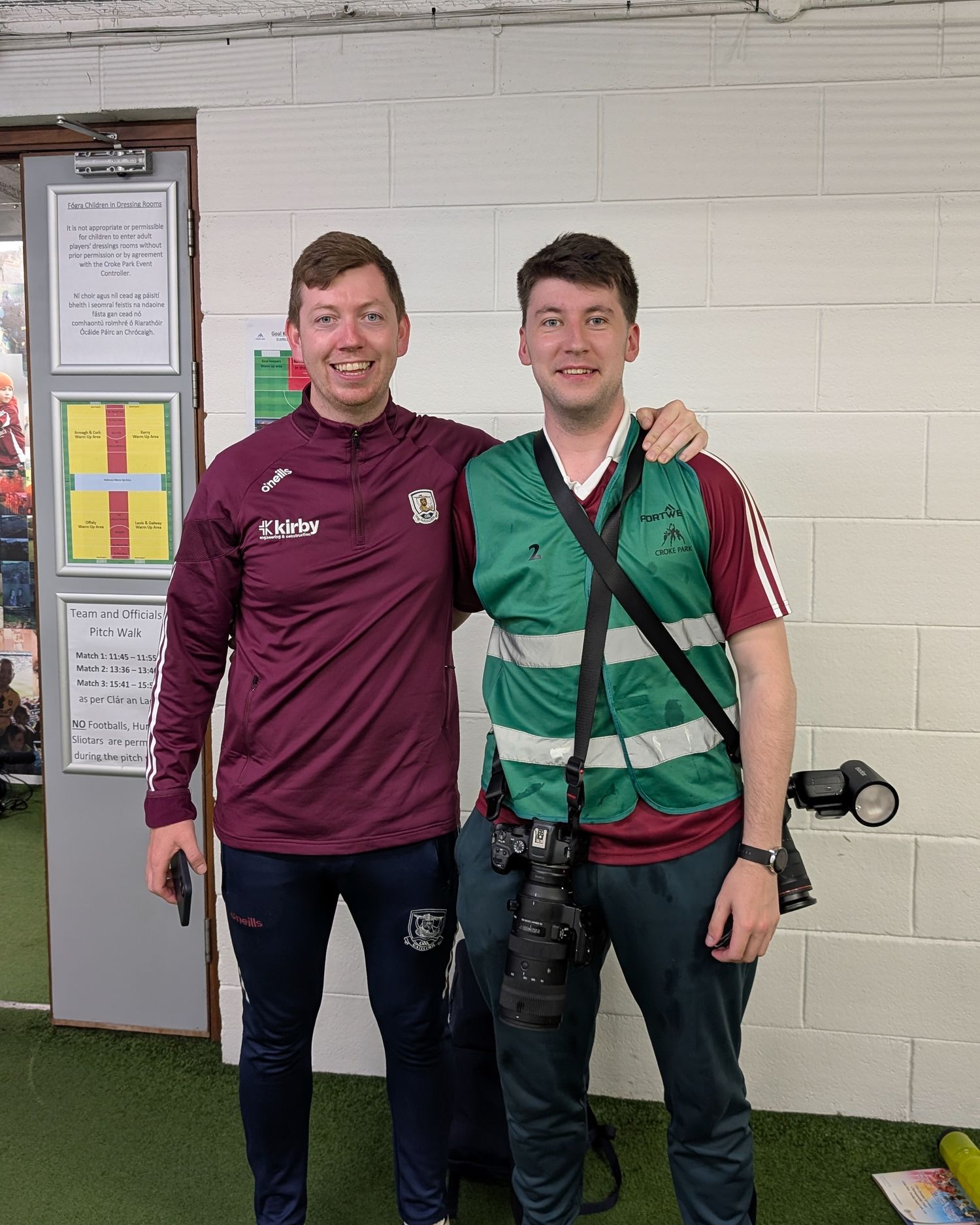 Two people wearing sports attire pose together indoors, one in maroon and one in green with a camera.