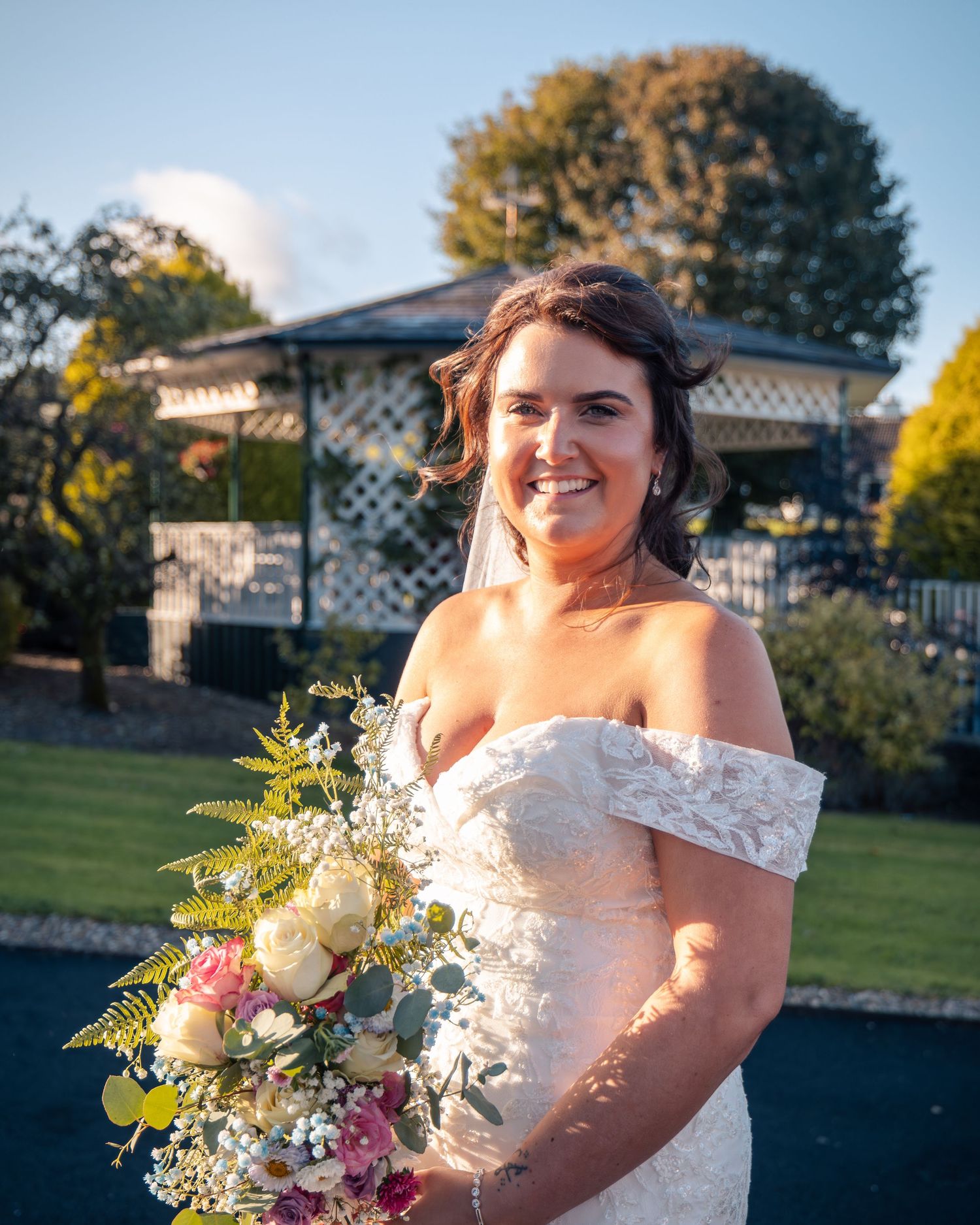 Bride in off-shoulder wedding dress holds colorful bouquet in outdoor sunlit setting.