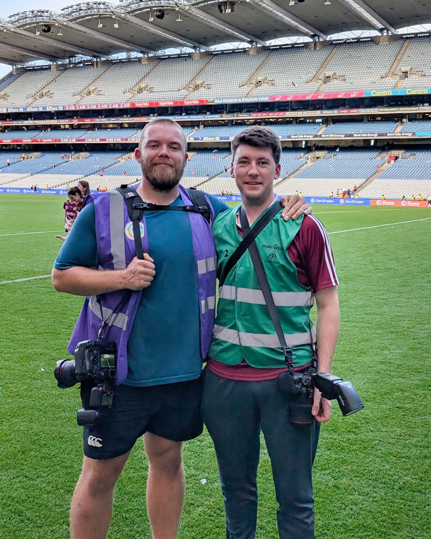 Two sports photographers wearing matching green jerseys stand together on a stadium field with camera equipment.