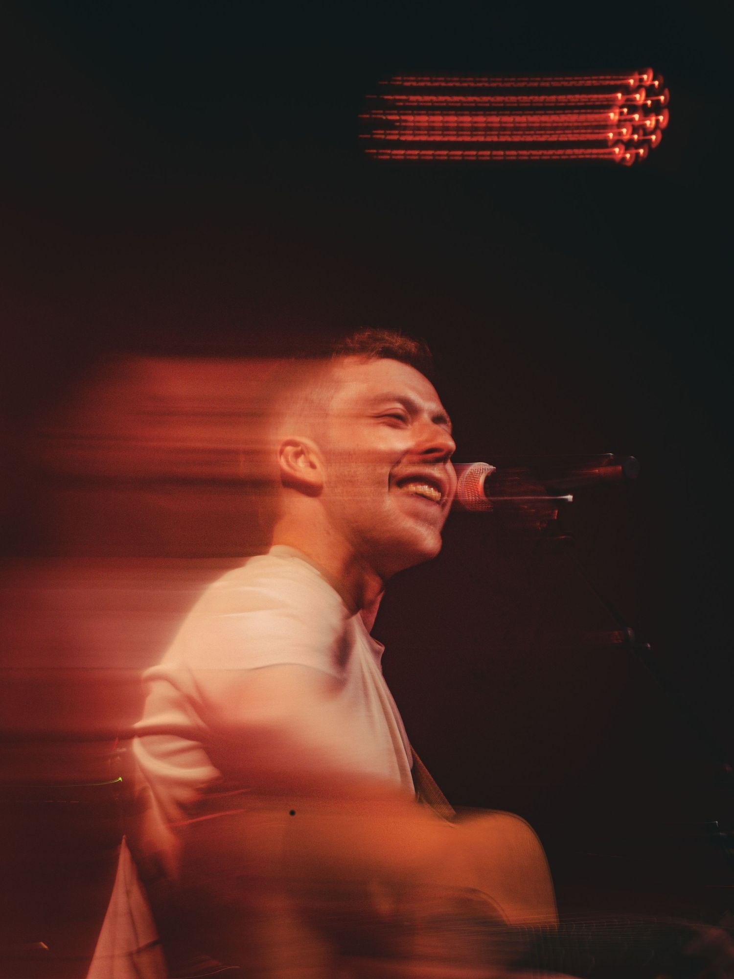 A performer in a white shirt creates a motion blur effect while singing on stage under dramatic red lighting.