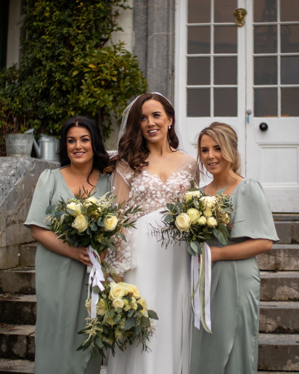 Bridesmaids in sage green dresses hold elegant white and cream floral bouquets while posing outside a historic building.