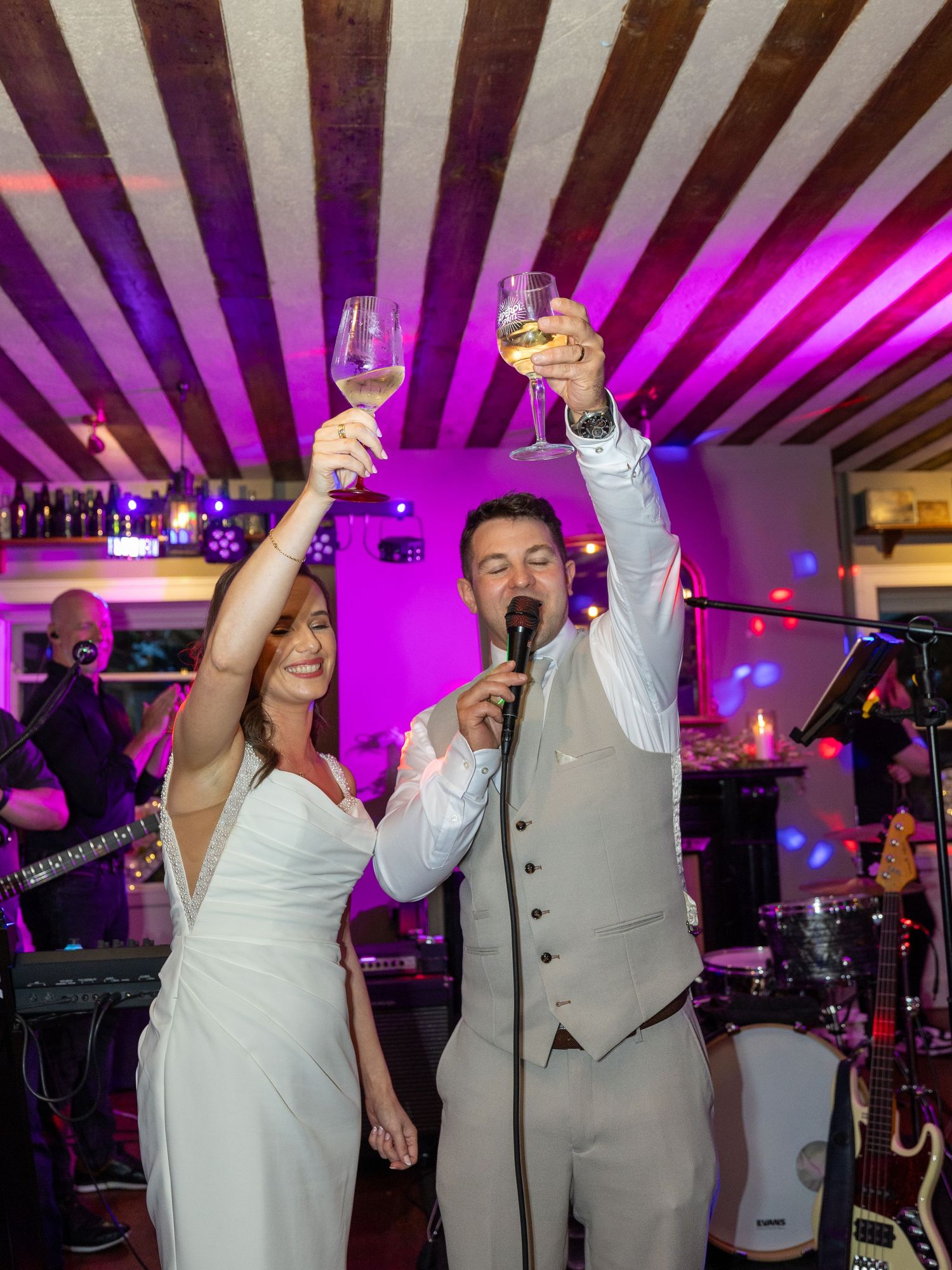 Wedding celebration with couple and guests raising glasses under purple lights and striped ceiling.