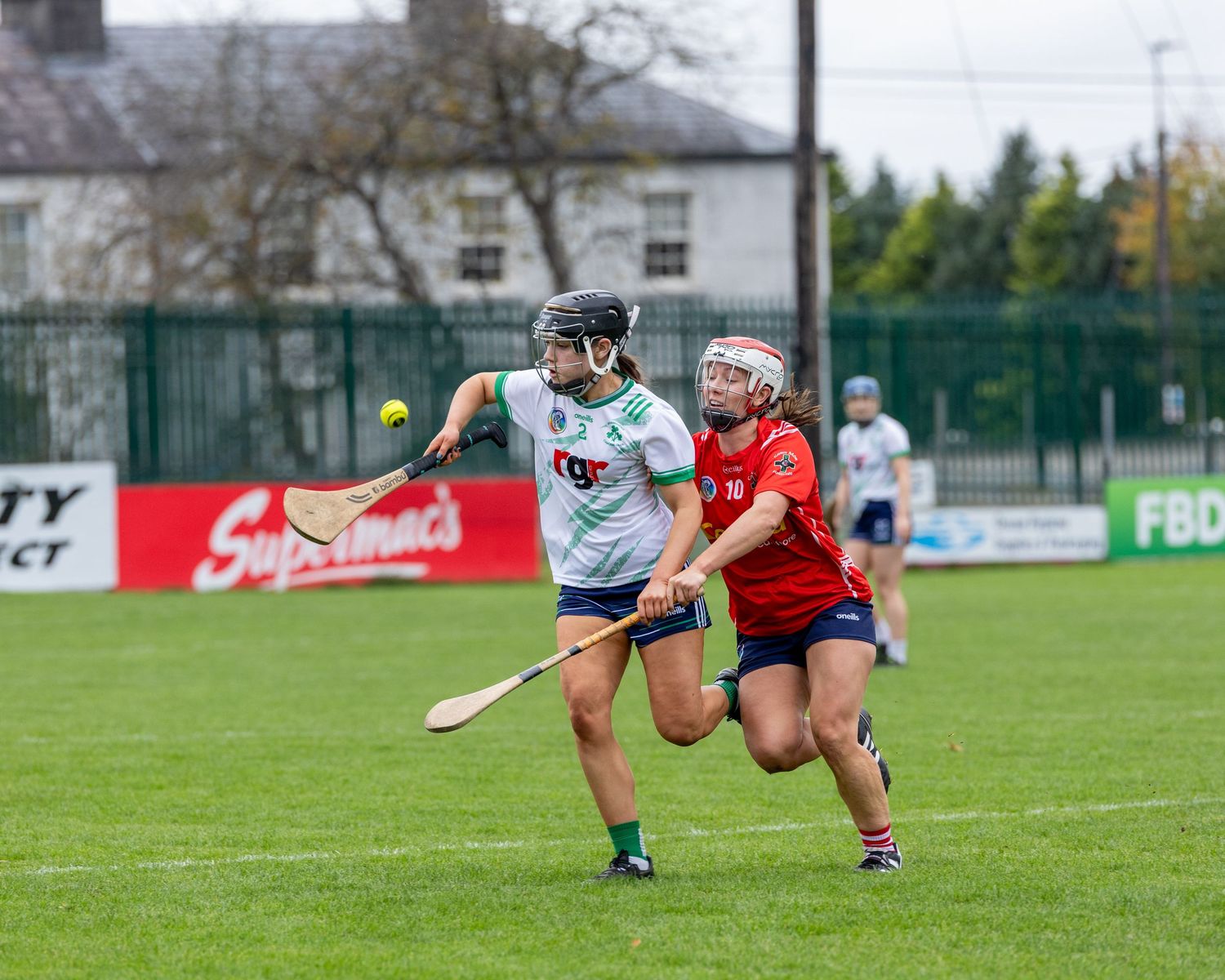 Two camogie players compete for possession during a match on a grass pitch with advertising boards visible in background.