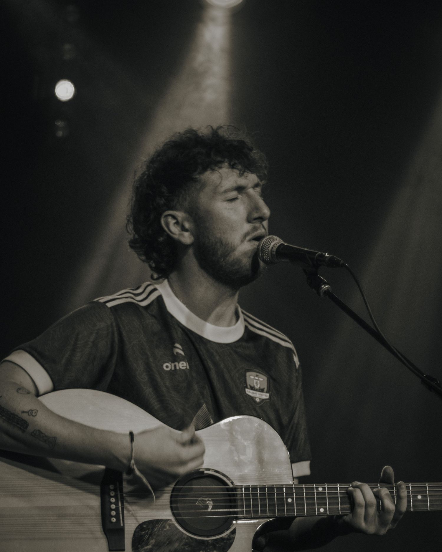 Musician performing with an acoustic guitar under dramatic stage lighting in black and white.