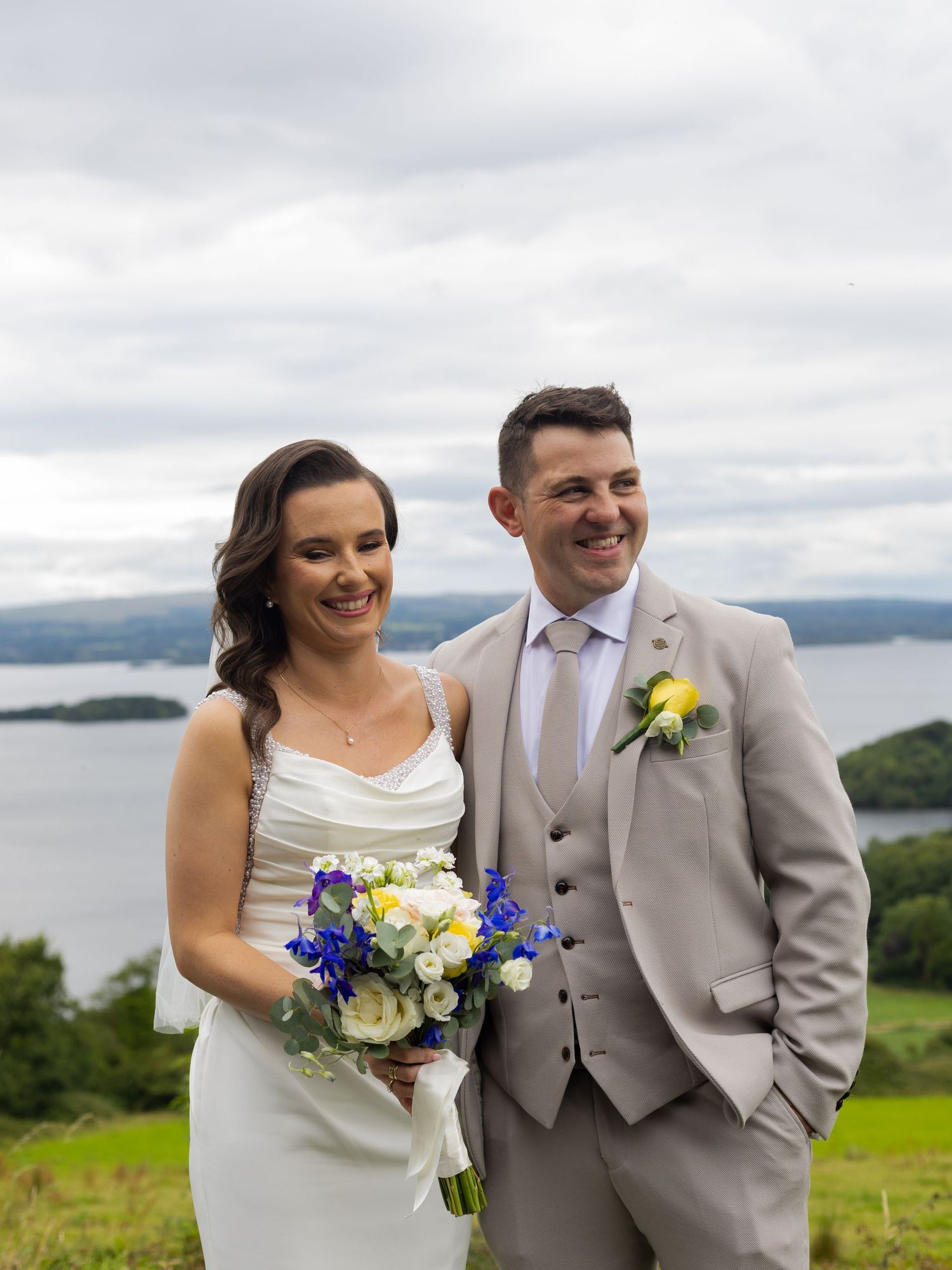 Wedding couple poses with bouquet of blue and white flowers against scenic coastal mountain backdrop.