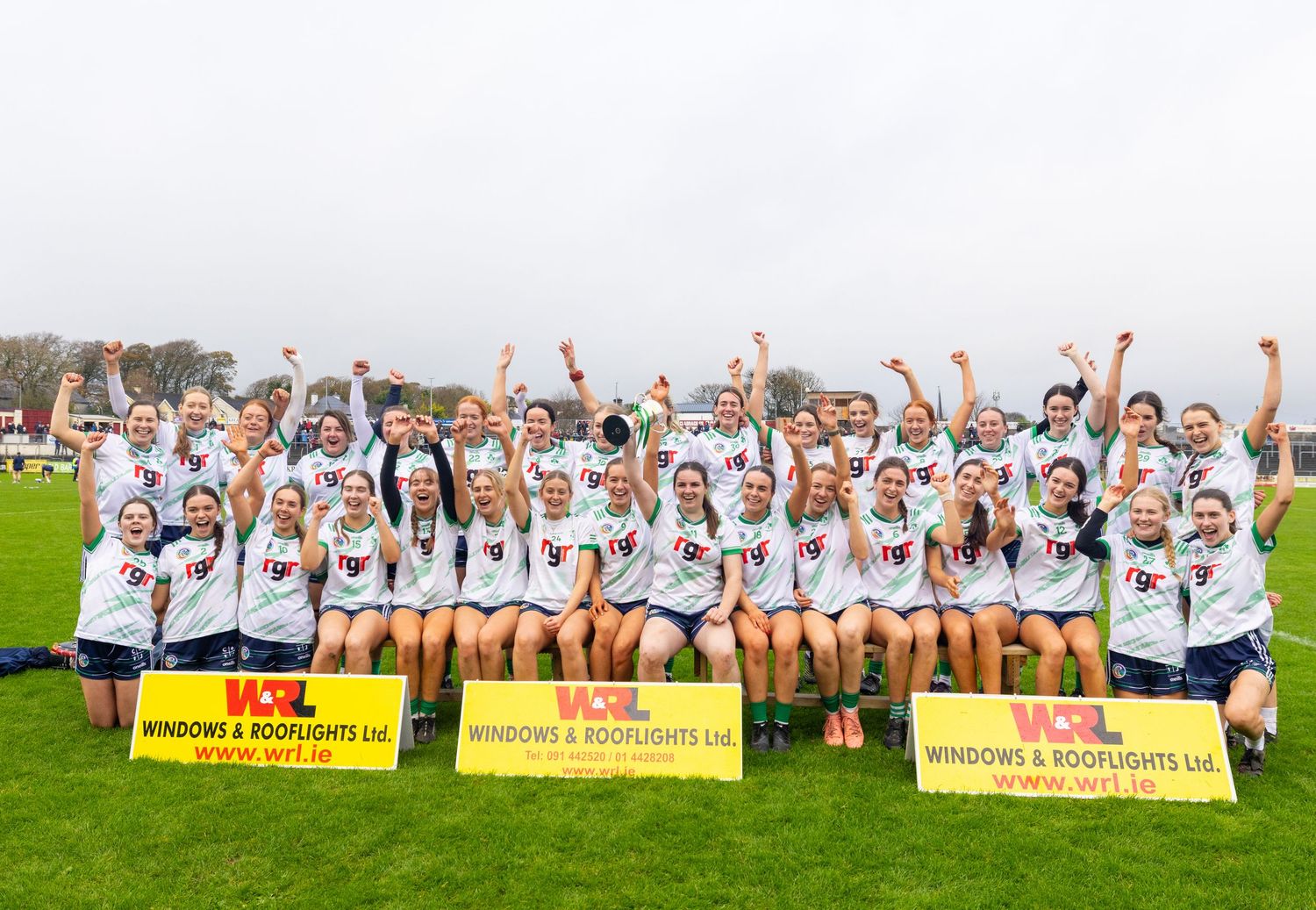 A group of athletes in white uniforms celebrate together on a sunny field during a sports event.