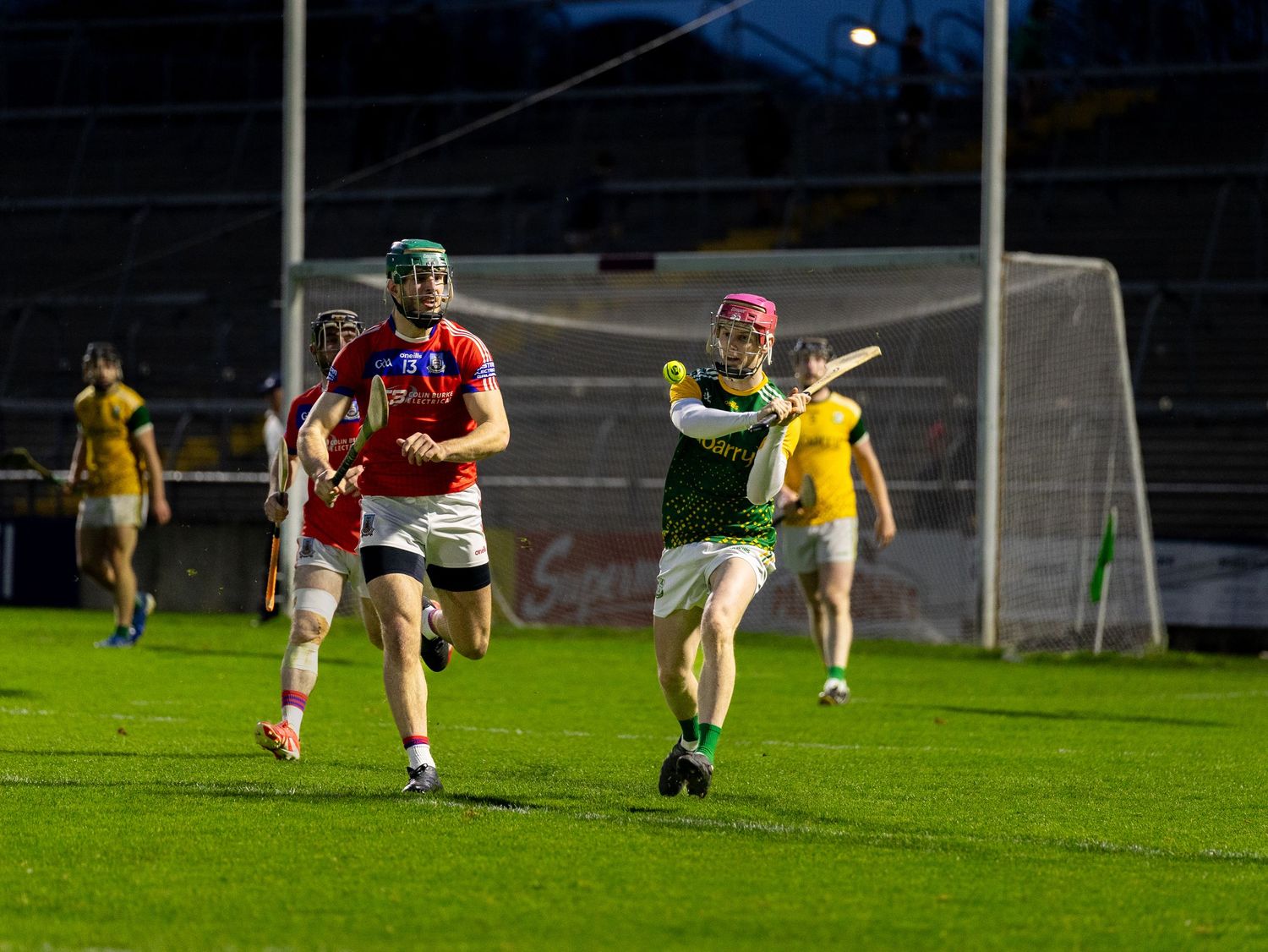 A Gaelic football match in progress at night with players in red and yellow jerseys competing on a floodlit field.