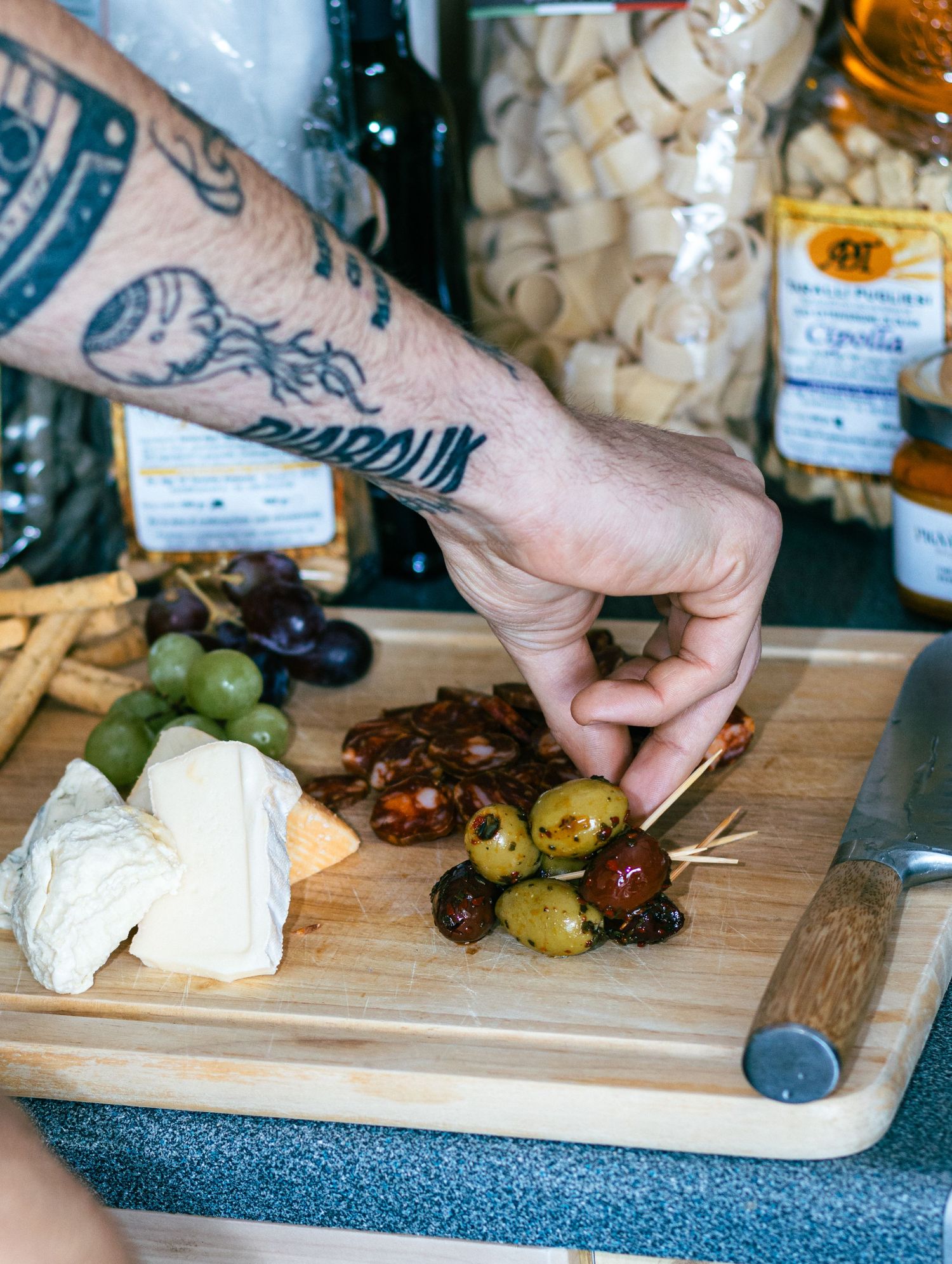Hands arranging cheese, olives and grapes on a wooden charcuterie board.