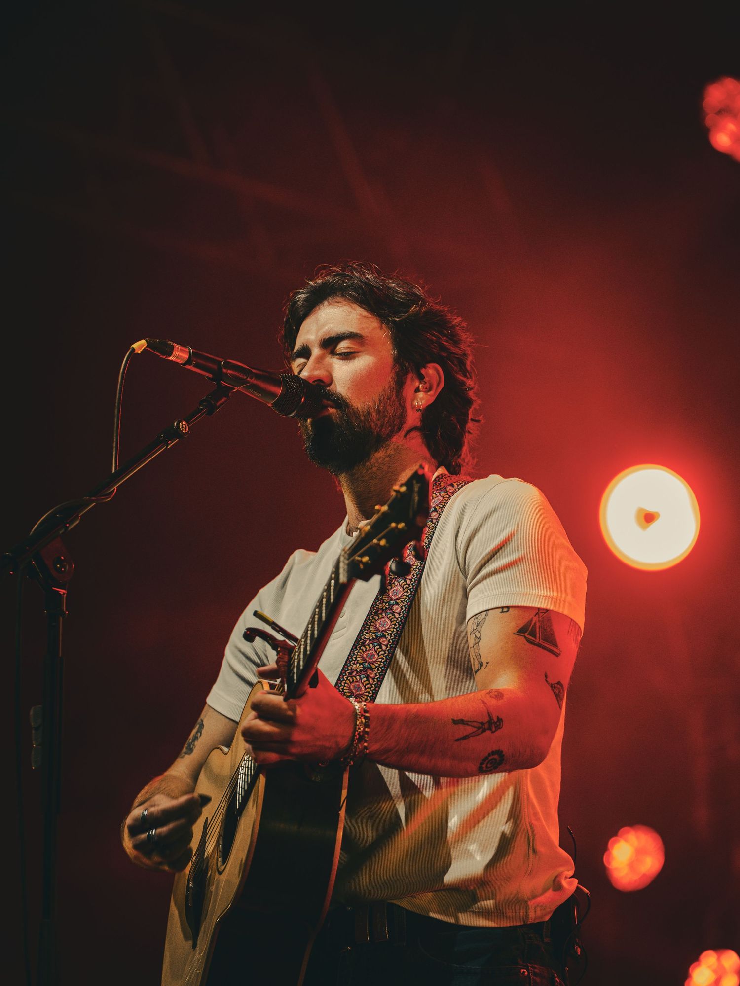 Concert performer singing into microphone under dramatic red stage lighting during live performance.