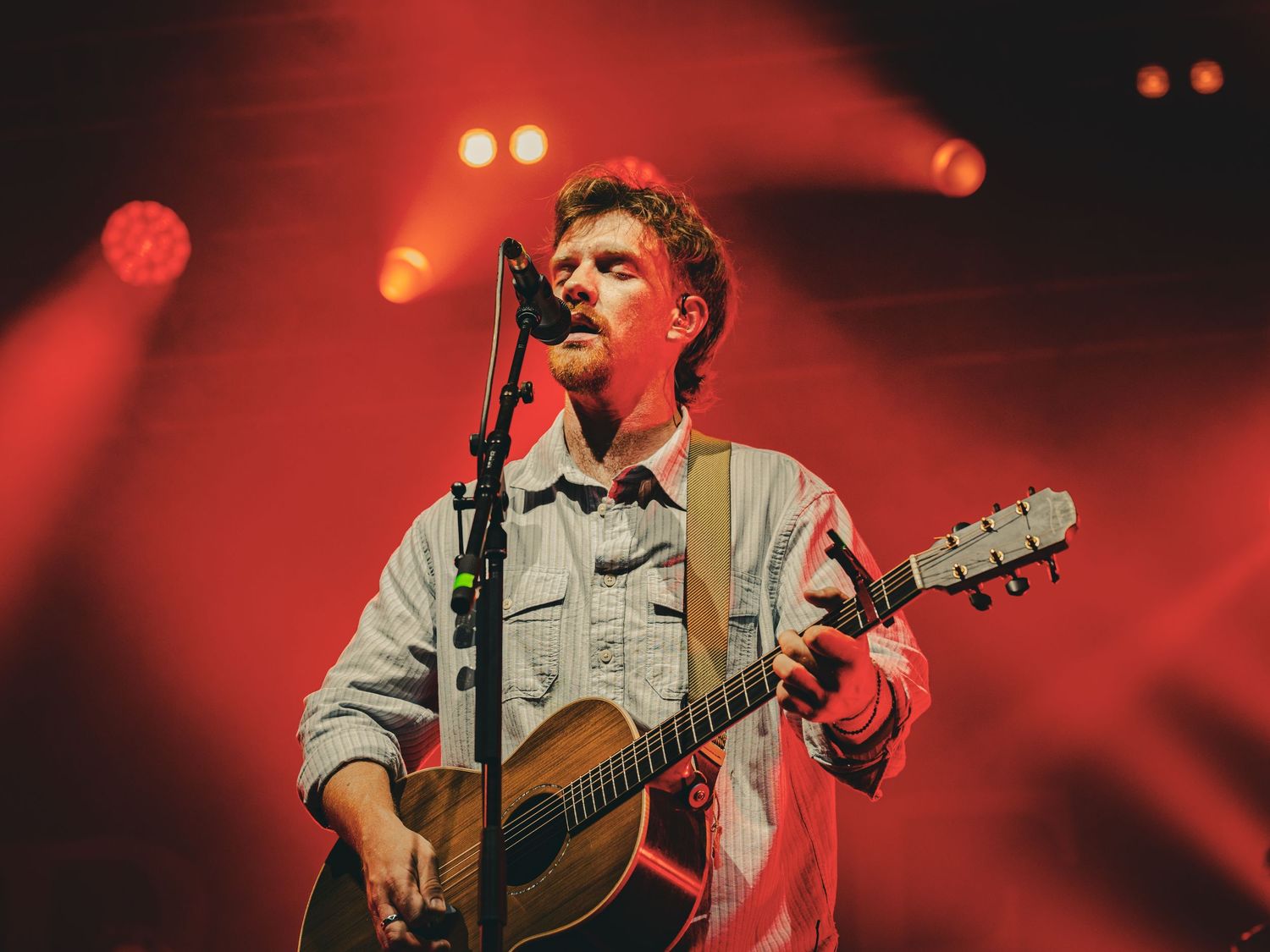 A performer plays acoustic guitar on stage with dramatic red lighting and smoke effects in the background.