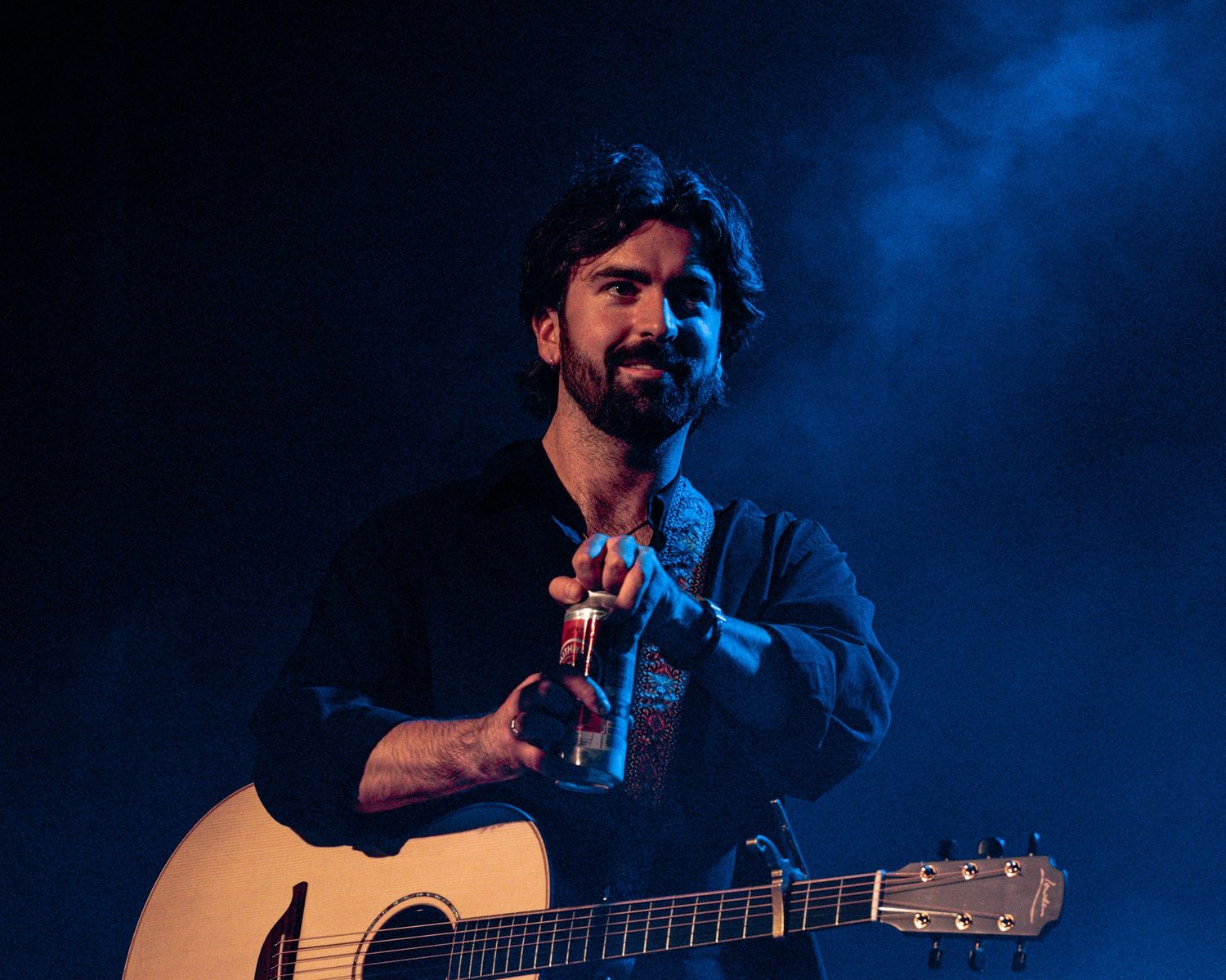 A musician performs on stage with an acoustic guitar against a dramatic blue-lit backdrop.