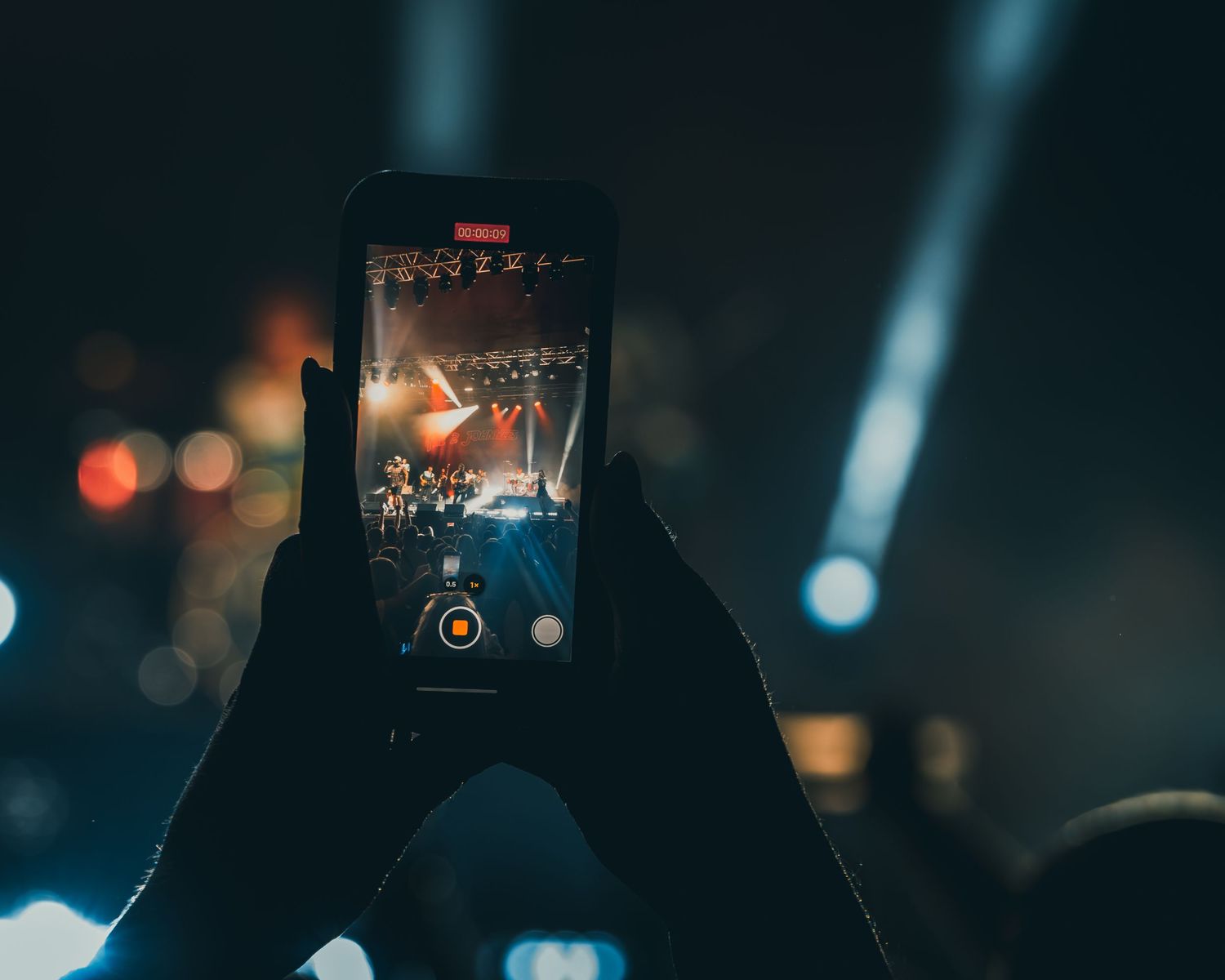 Smartphone capturing concert stage performance through screen with blurred lights in background.