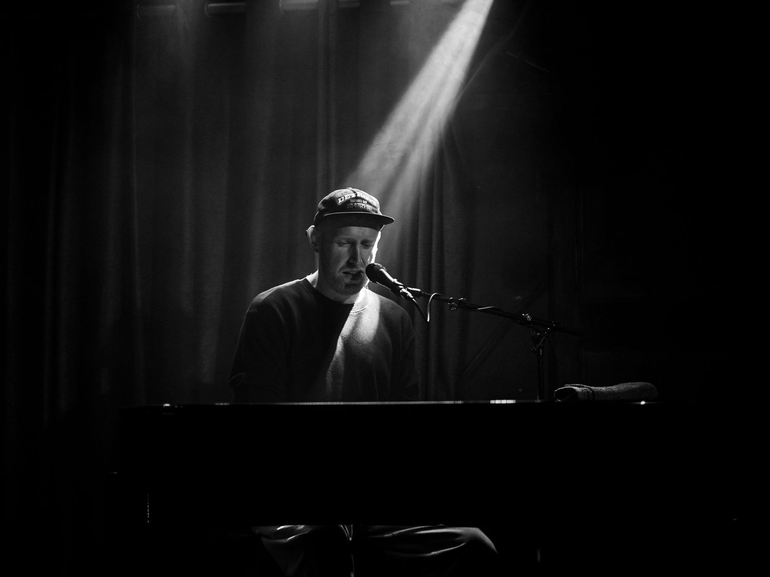 Dramatic black and white stage photo of a musician performing at a piano under a spotlight in darkness.